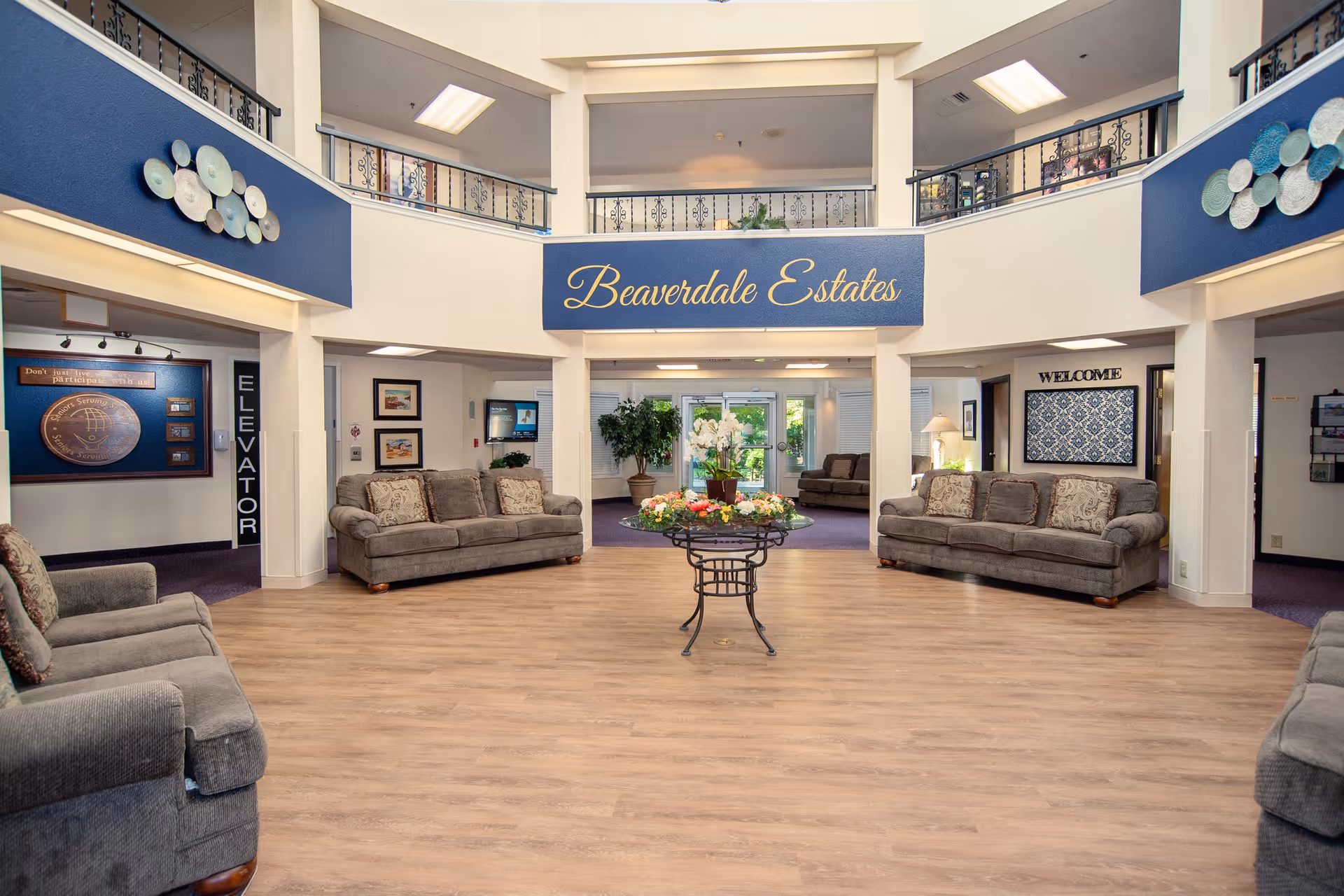 Spacious and well-lit senior living facility lobby with multiple gray sofas arranged around the room, a round table with a floral centerpiece in the center, and a sign reading 'Beaverdale Estates' above the entrance. The area features wood flooring, blue accent walls with decorative plates, and a second-floor balcony with black railings.
