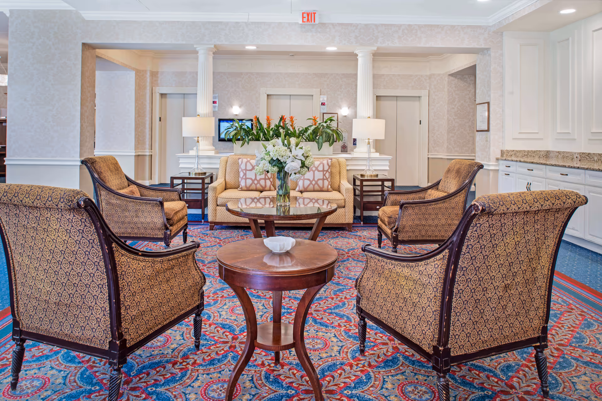 A well-decorated sitting area in a senior living facility with four patterned armchairs arranged around a small wooden table with a decorative bowl. Behind the chairs is a beige sofa with patterned cushions, two side tables with lamps, and a glass coffee table with a vase of flowers. The room features a patterned carpet, white columns, and light-colored wallpaper. Two elevators are visible in the background.