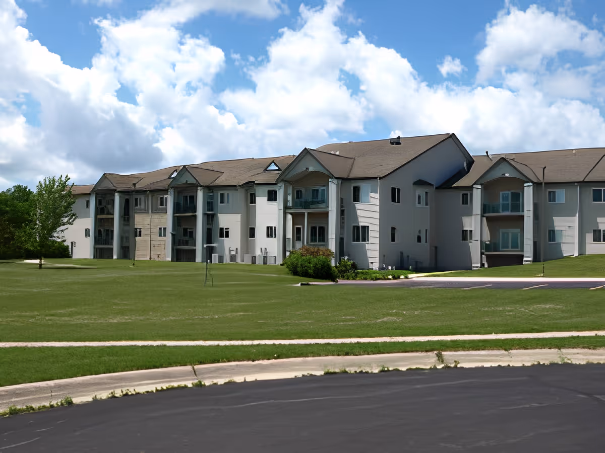 Exterior view of a large multi-story residential building with balconies, surrounded by a grassy lawn and a paved driveway under a partly cloudy blue sky.