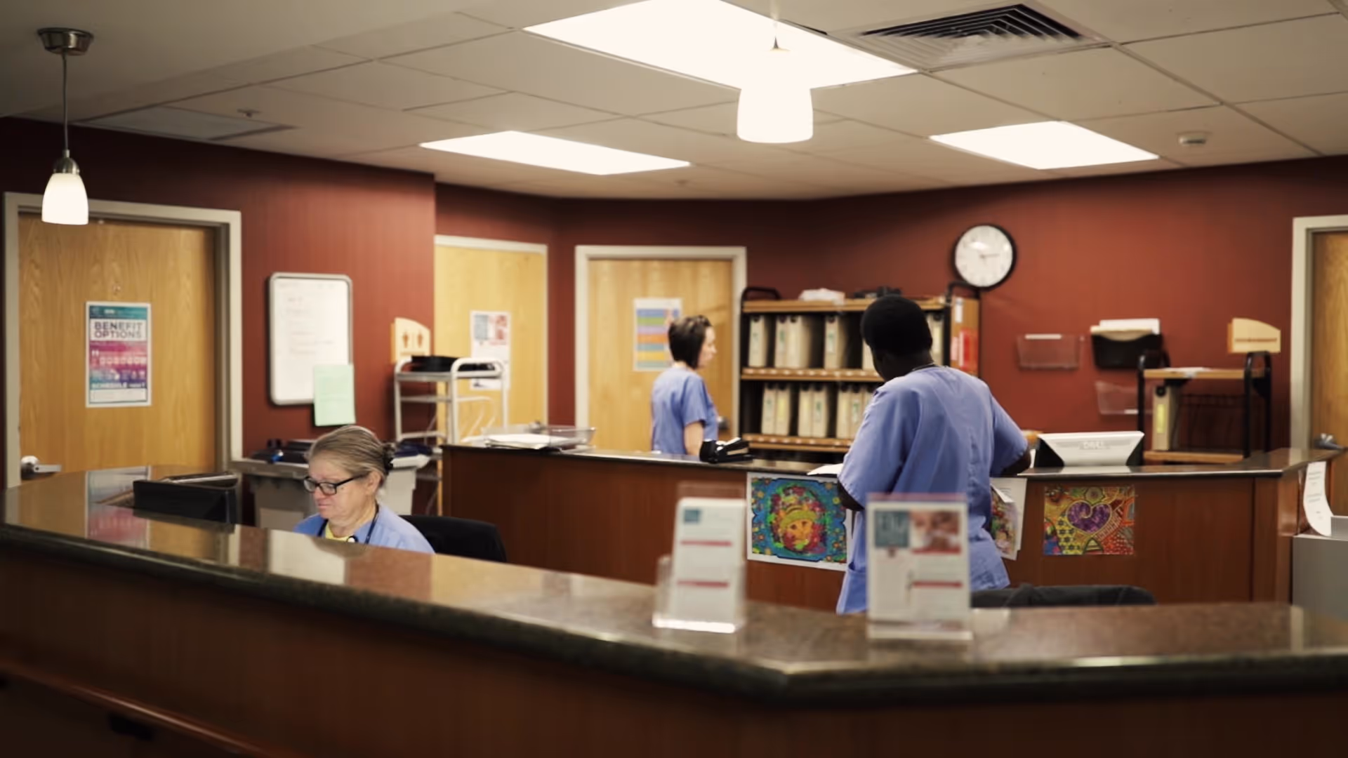 Reception area in a senior living facility with three staff members wearing scrubs working behind a curved counter. The background shows red walls, wooden doors, a clock, and shelves with organized files.