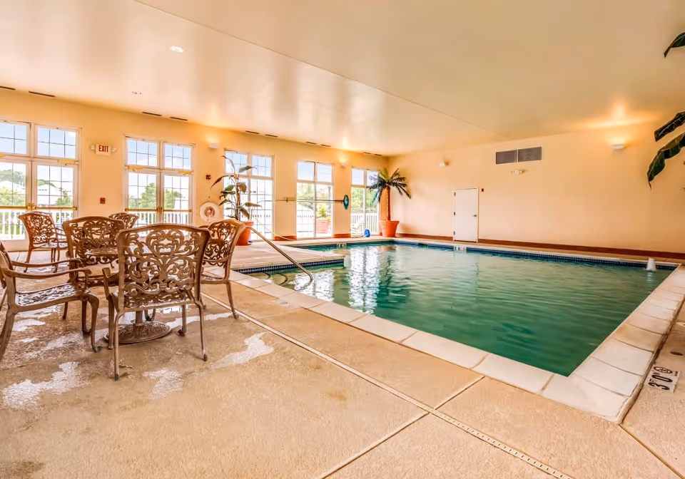 Indoor swimming pool area with clear water, surrounded by beige tiled flooring. There are ornate metal chairs and tables on the left side near large windows letting in natural light. Potted plants are placed around the pool area, and the walls are painted light beige.
