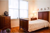 A cozy bedroom with a wooden bed frame and matching wooden dresser and nightstand. The bed is made with a light-colored quilt, and a lamp is turned on on the nightstand. There is a large window with white blinds letting in natural light, and decorative items are placed on the dresser and above the headboard.