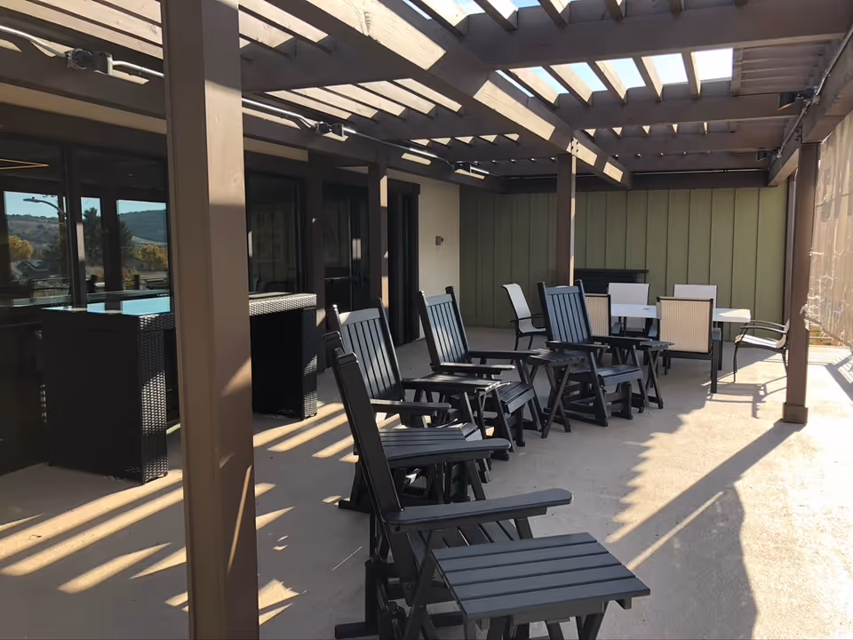 Outdoor patio area with several black wooden rocking chairs and a table with chairs under a pergola structure. The patio is adjacent to a building with large windows and doors, and there is a scenic view of hills in the background.