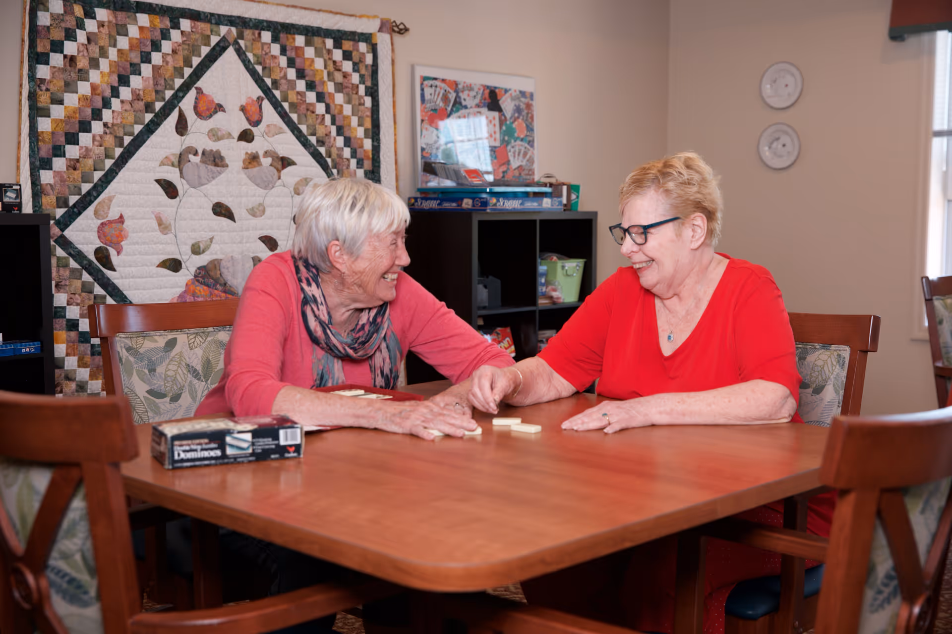 Two elderly women sitting at a wooden table playing dominoes and smiling at each other in a cozy room with a quilt hanging on the wall and shelves with board games in the background.