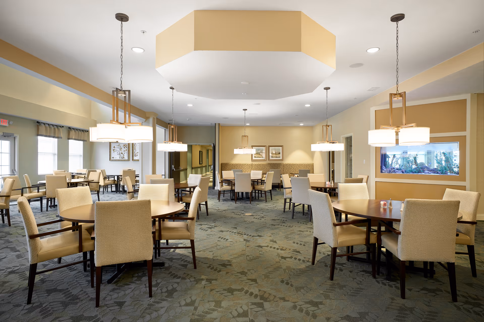A spacious dining room with multiple round tables surrounded by beige upholstered chairs. The room features modern pendant lights hanging from the ceiling, a patterned carpet, and a large built-in aquarium on one wall. The walls are painted in soft yellow and beige tones, and there are framed pictures and windows letting in natural light.