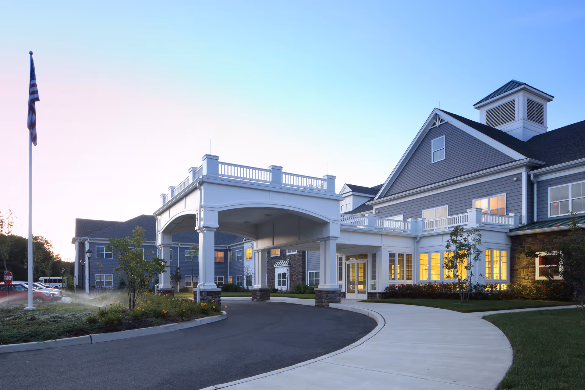 Exterior view of Sunnyside Manor senior living facility at dusk, showing a large covered entrance with white pillars, a curved driveway, well-maintained landscaping, and an American flag on a flagpole.