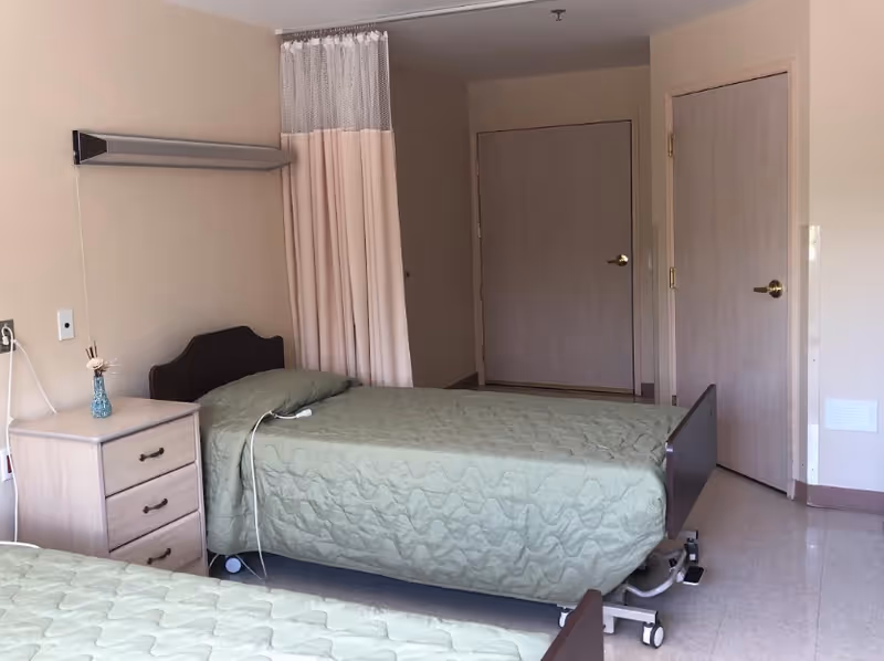 Patient room in a skilled nursing facility with two hospital-style beds, a nightstand, a privacy curtain, and closed doors.