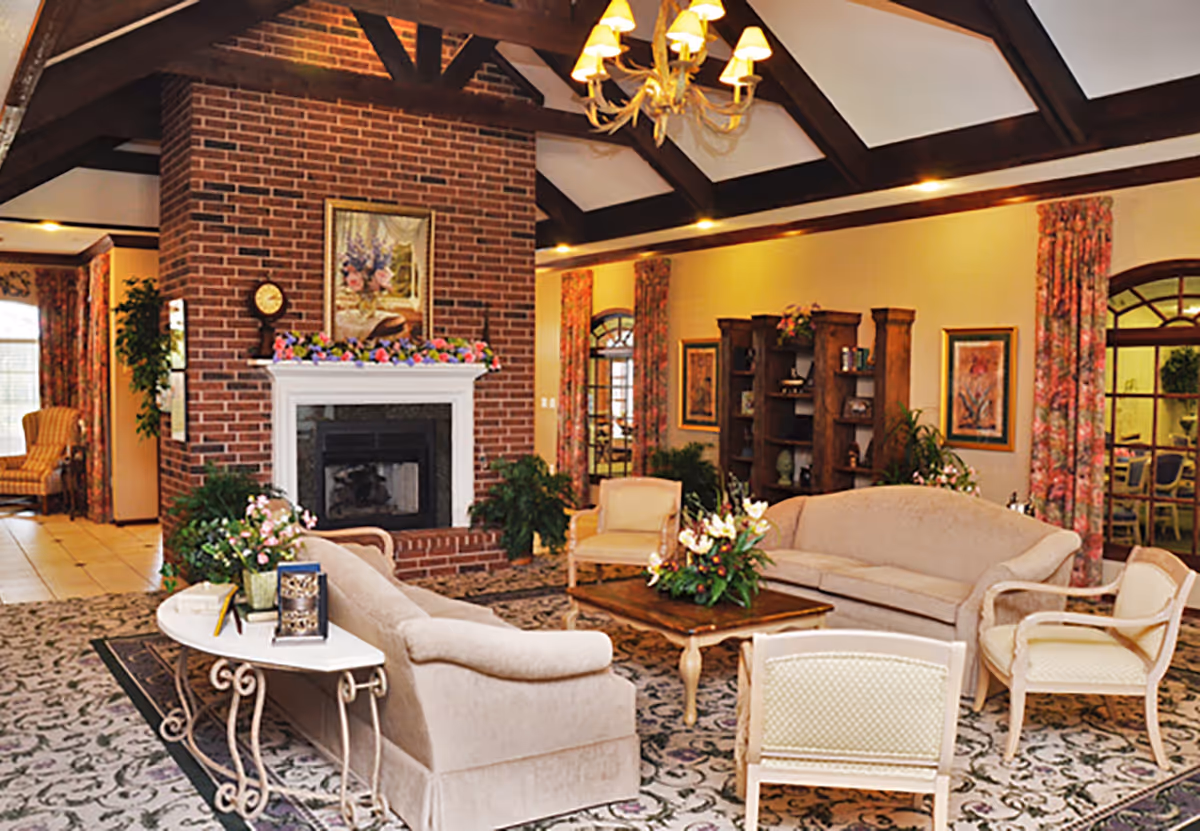 A cozy common lounge with sofas and armchairs arranged around a brick fireplace beneath exposed wooden ceiling beams and a chandelier.