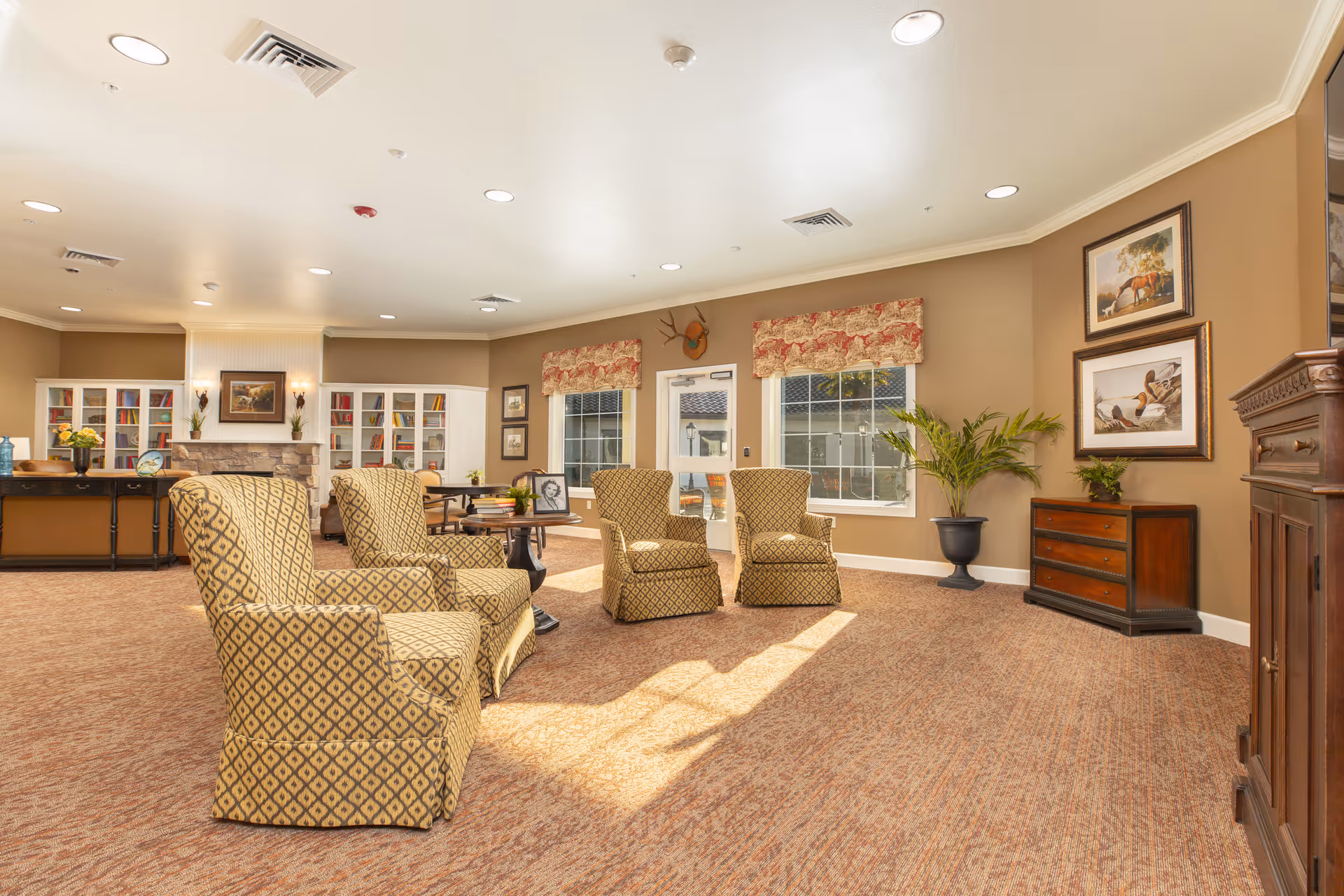 A bright and spacious living room area with four patterned armchairs arranged around a small table. The room features beige walls, carpeted flooring, large windows with floral valances, framed artwork, a potted plant, and built-in white bookshelves with a fireplace in the background.