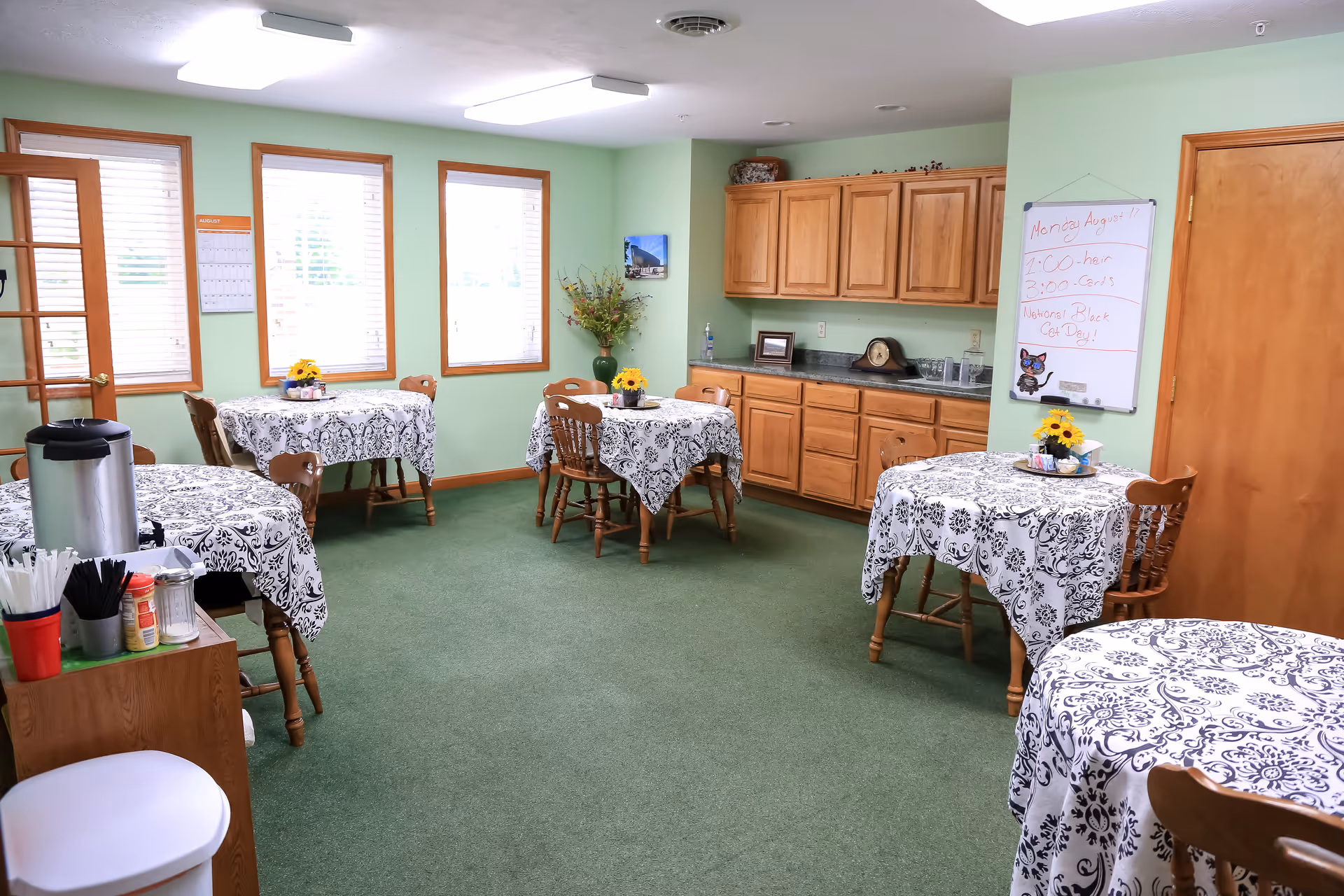 Small dining/activity room with round tables covered in patterned tablecloths, wooden chairs, a kitchenette along the back wall, and green carpet.