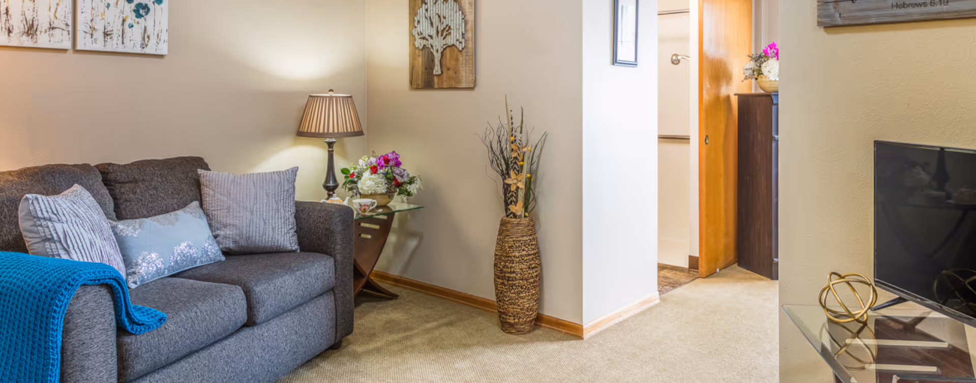 A cozy living room area with a gray sofa adorned with multiple pillows and a blue throw blanket. Next to the sofa is a side table with a lamp and a floral arrangement. The walls are decorated with framed artwork and a wooden wall hanging. A tall vase with decorative branches is placed near the corner. A flat-screen TV is visible on a glass stand to the right, and a hallway leads to another room.