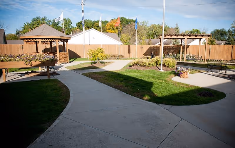 Outdoor courtyard area with paved walkways, green grass, flower beds, wooden gazebos, benches, and several flags on poles against a backdrop of trees and a wooden fence.