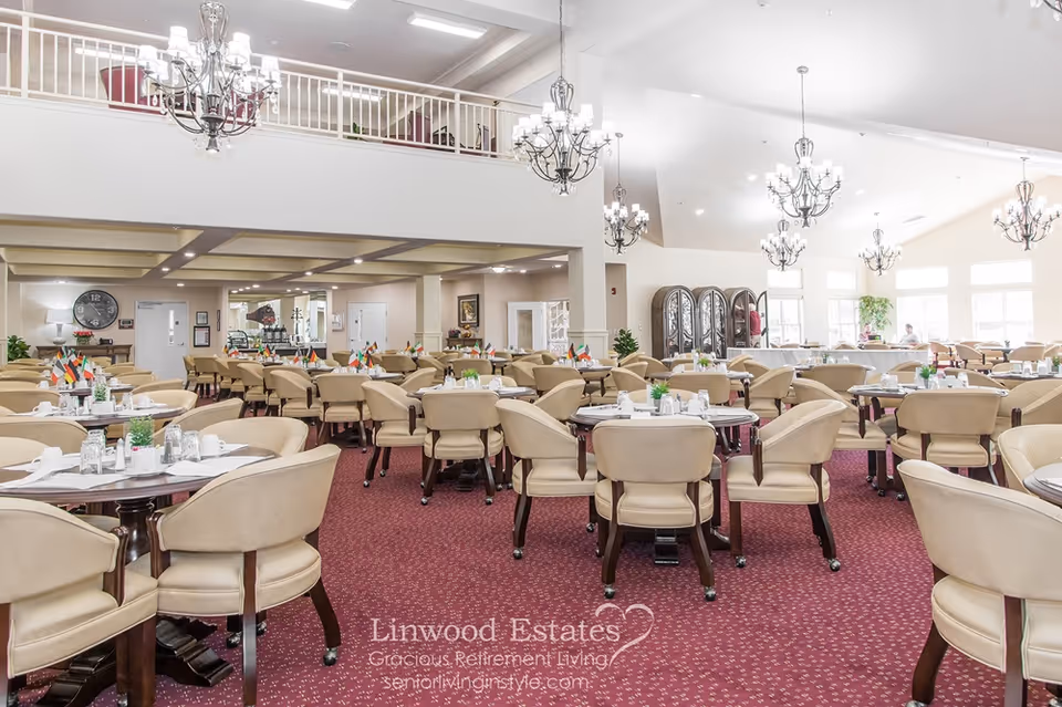 Large bright dining room in a senior living facility with many round tables, beige chairs, and chandeliers.
