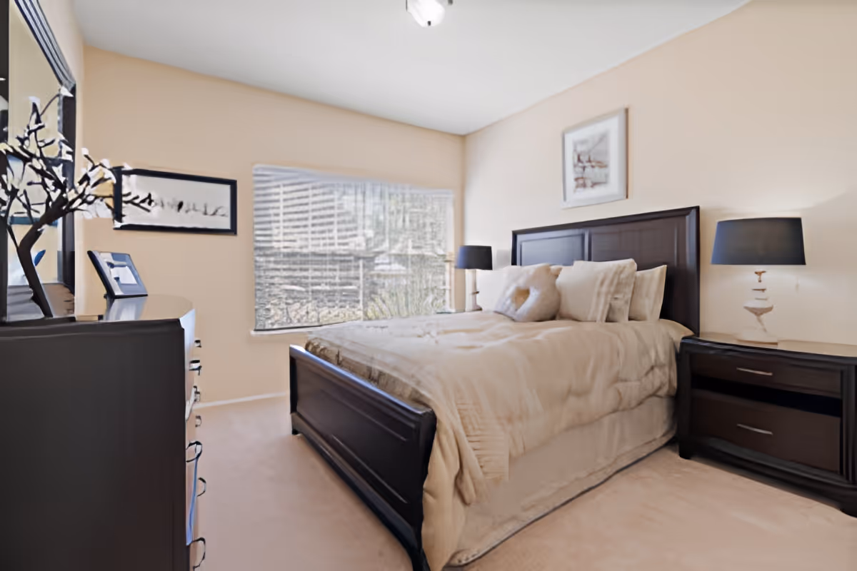 A cozy bedroom with a large bed featuring beige bedding and multiple pillows. The room has light beige walls, a window with blinds letting in natural light, two dark wooden nightstands with matching lamps, a dresser with decorative items, and framed artwork on the walls.