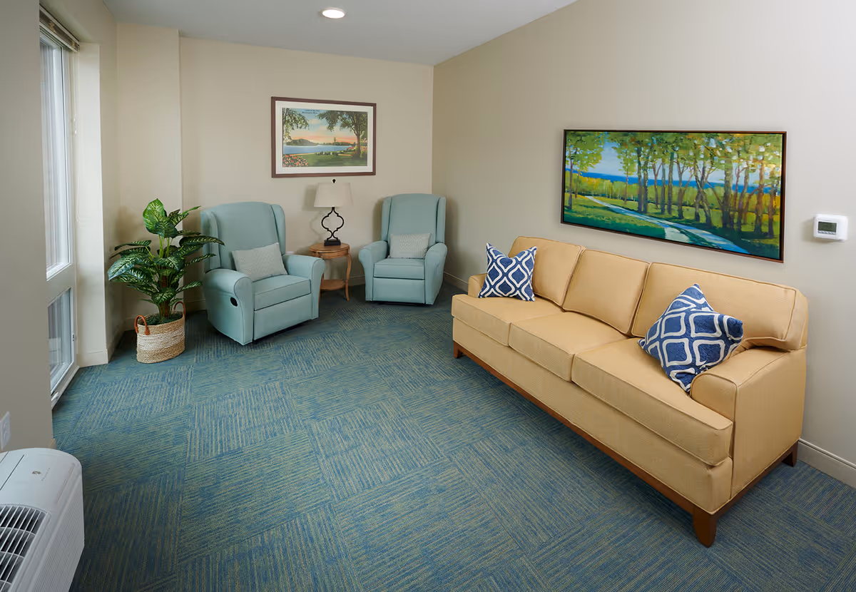 Comfortable sitting room with a beige sofa, two light-blue armchairs, a side table and lamp, artwork on the walls, and a potted plant.