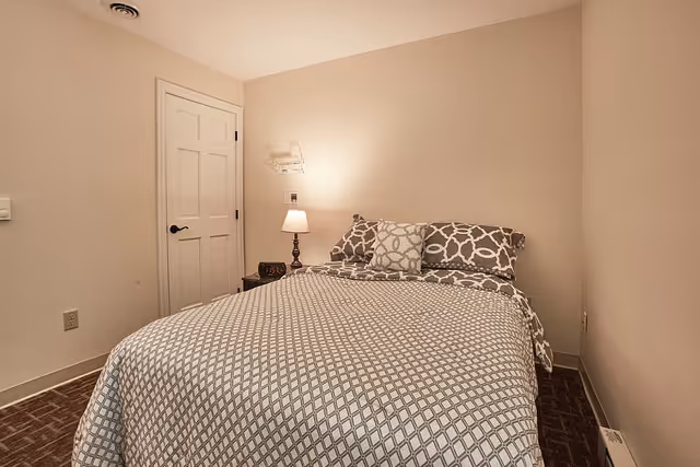 A small bedroom with a bed covered in a patterned gray and white bedspread and matching pillows. There is a nightstand with a lamp and a digital clock next to the bed. The walls are beige, and there is a closed white door on the left side of the room.