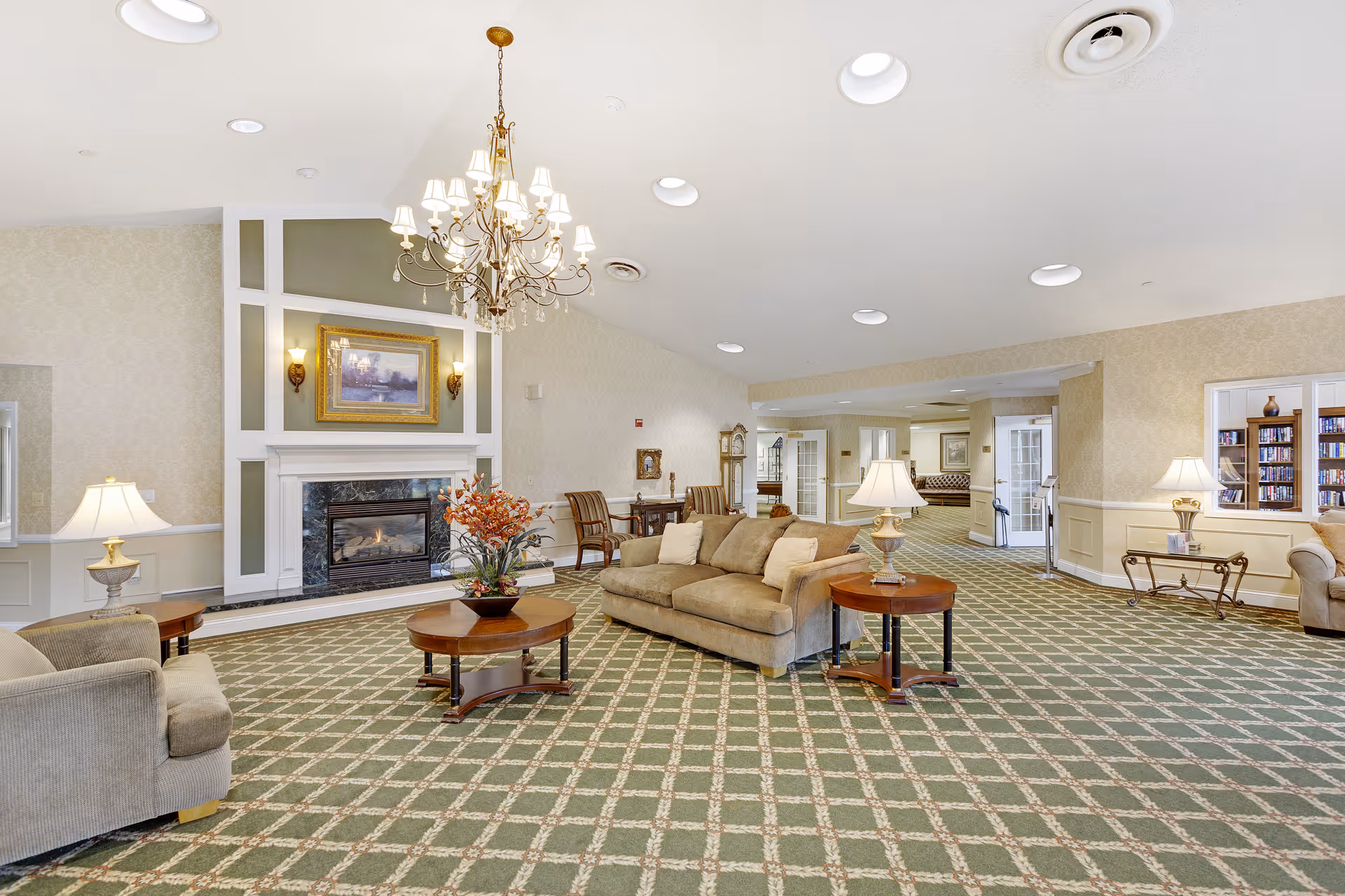 A spacious and well-lit living room area in a senior living facility featuring a patterned green carpet, beige sofas with cushions, wooden coffee and side tables with lamps, a decorative chandelier, and a fireplace with a framed painting above it. The room has light-colored walls with wainscoting and built-in bookshelves, creating a cozy and inviting atmosphere.