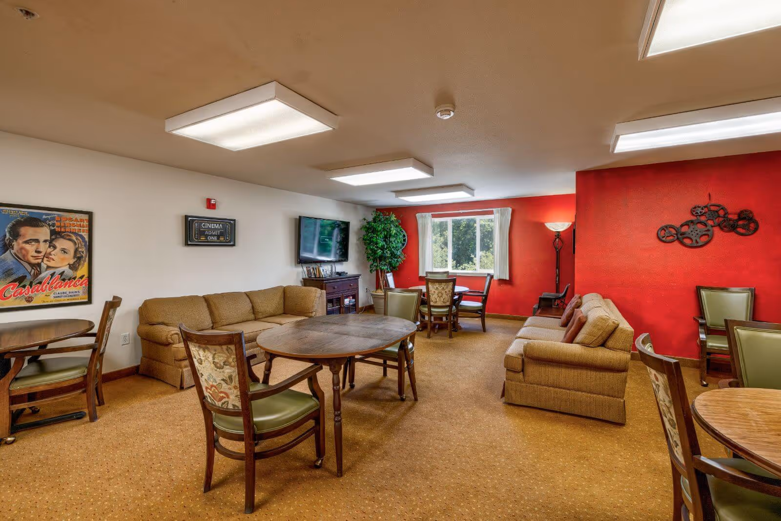 A cozy common area with beige sofas, wooden tables, and chairs. The room has a red accent wall with decorative film reel art, a large window with white curtains, a flat-screen TV mounted on the wall, and a framed Casablanca movie poster. The ceiling has rectangular fluorescent lights, and there is a tall floor lamp near the window.