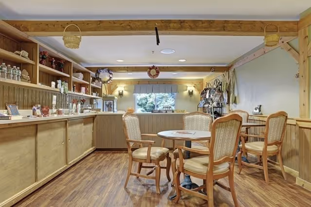 A cozy common area with wooden flooring and wooden furniture, including a round table surrounded by four cushioned chairs. The room features a long wooden counter with shelves above holding water bottles, cups, and decorative items. There are two wall sconces on the far wall, a window with a blue valance, and various hanging decorations and kitchen utensils on the walls.