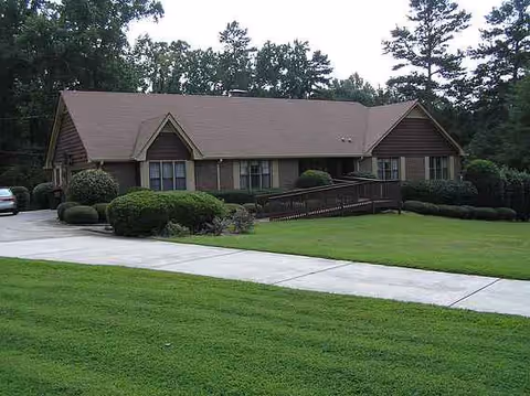 Single-story brown senior care home with a ramp, manicured lawn, and surrounding trees.