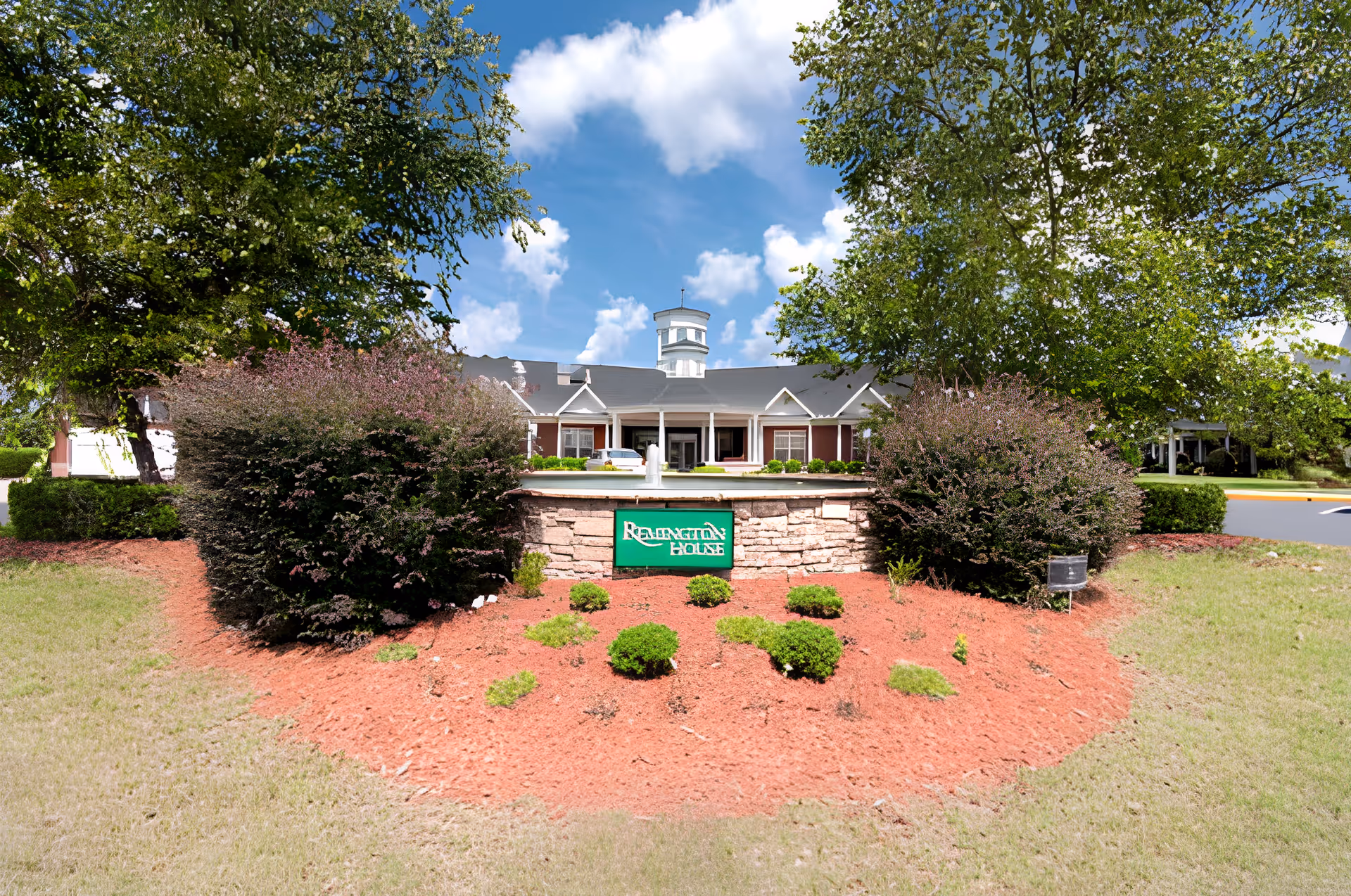 Front exterior view of a senior living facility named Remington House, featuring a landscaped garden with bushes and small plants, a stone sign with the facility's name, and a building with a cupola under a partly cloudy blue sky.