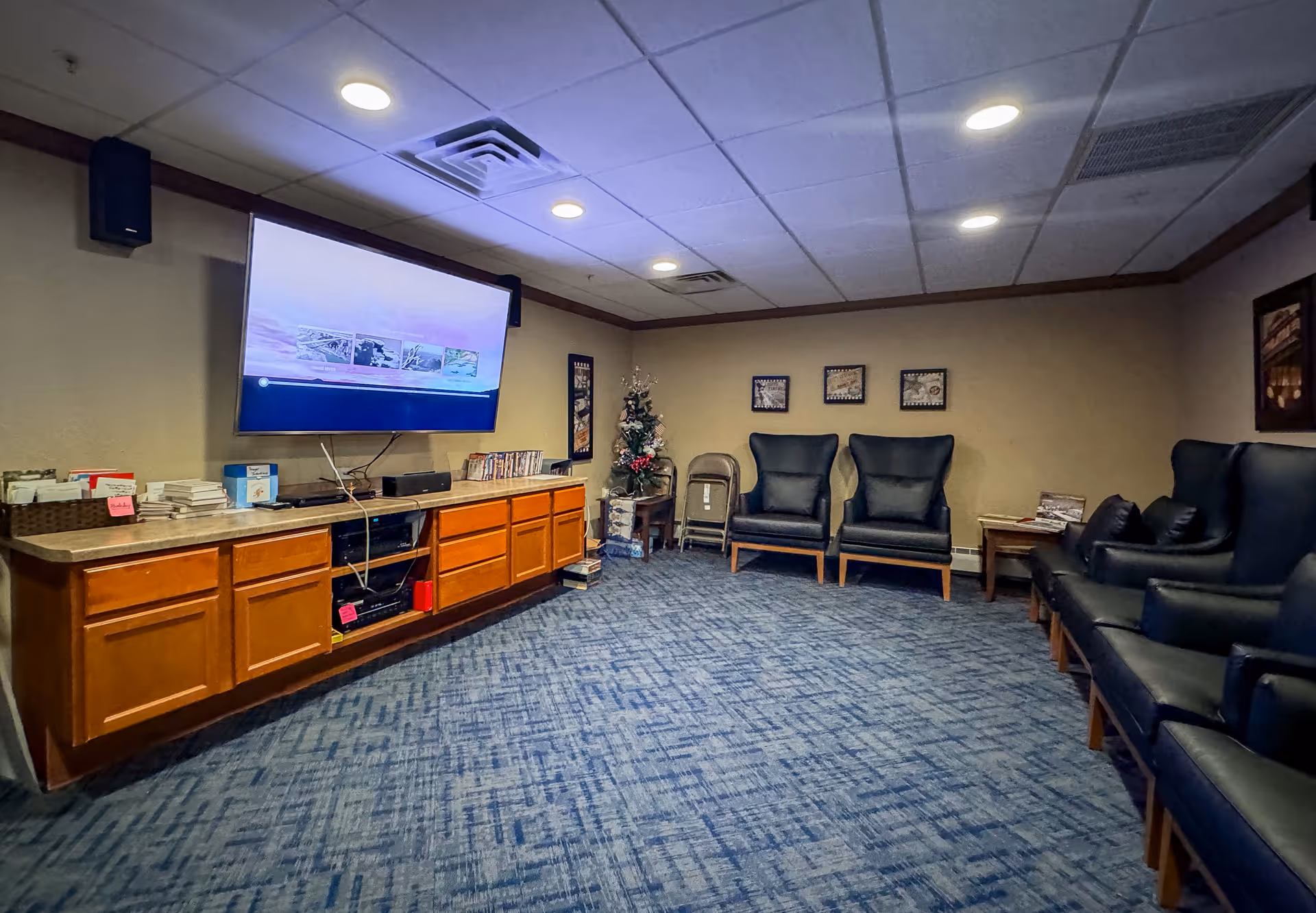 Carpeted lounge with a wall-mounted TV above a long wooden cabinet and several armchairs arranged along the walls.