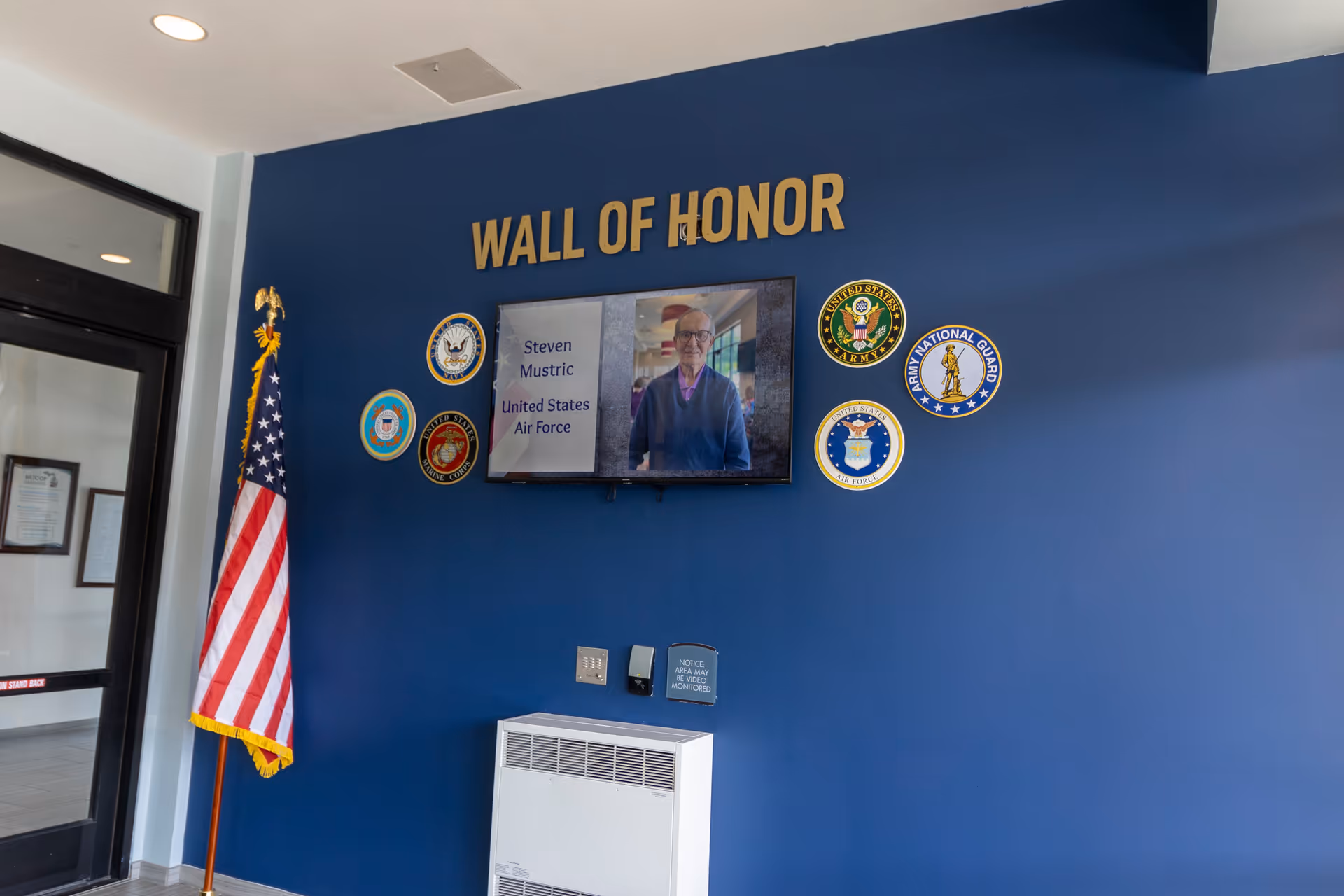 A blue lobby wall labeled "WALL OF HONOR" with military emblems, a mounted screen showing an honoree, and an American flag nearby.