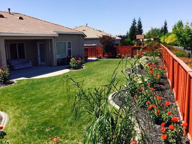 A well-maintained backyard garden area with green grass, a variety of plants and flowers along a red wooden fence, and a covered patio attached to a beige house with a tiled roof under a clear blue sky.