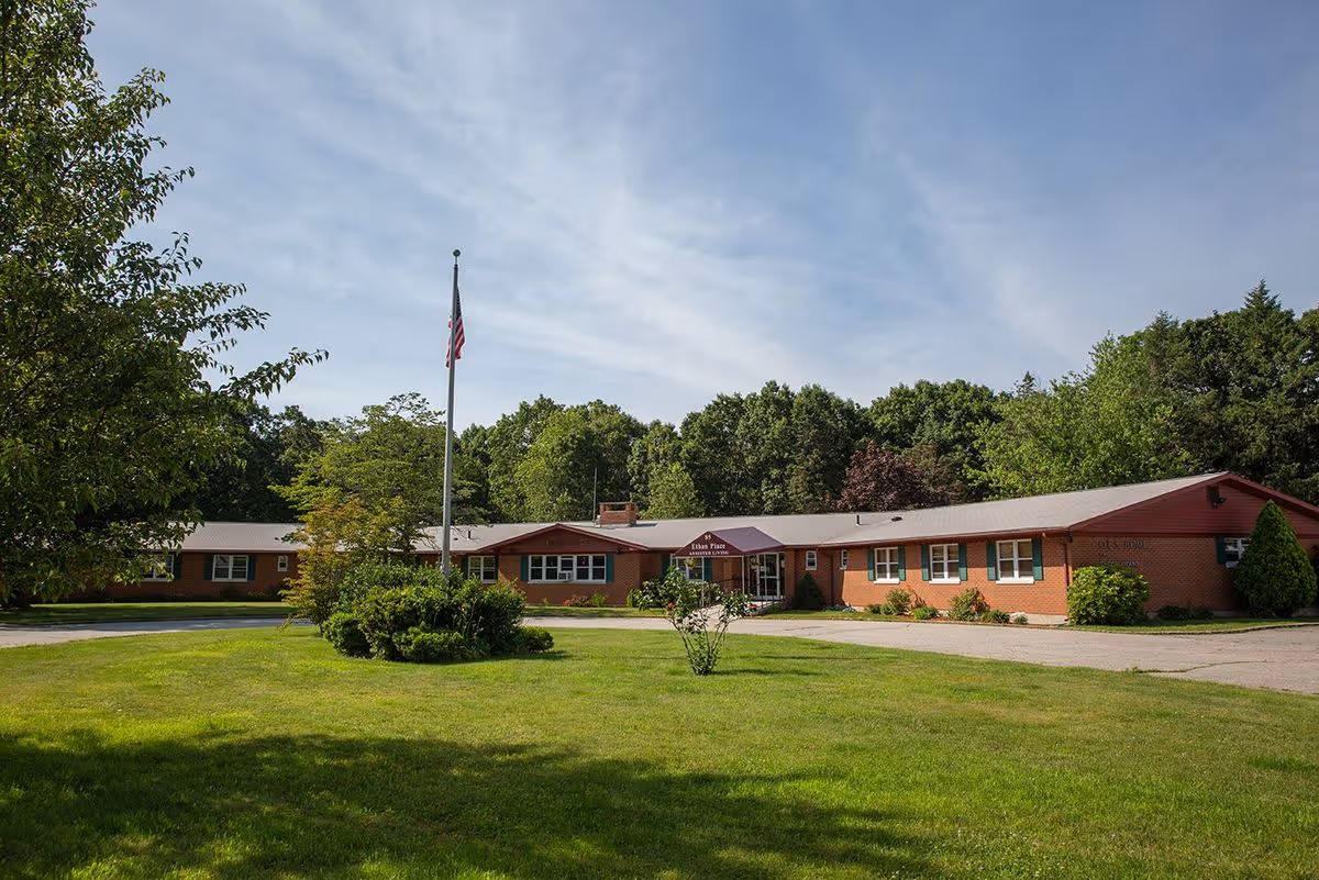 Single-story brick building labeled 'Ethan Place' with a front lawn, flagpole, and surrounding trees under a blue sky.