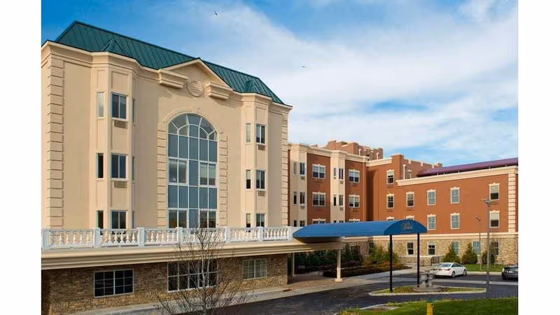 Front exterior of a multi-story beige and red-brick assisted living building with a large arched central window and a blue covered entrance.