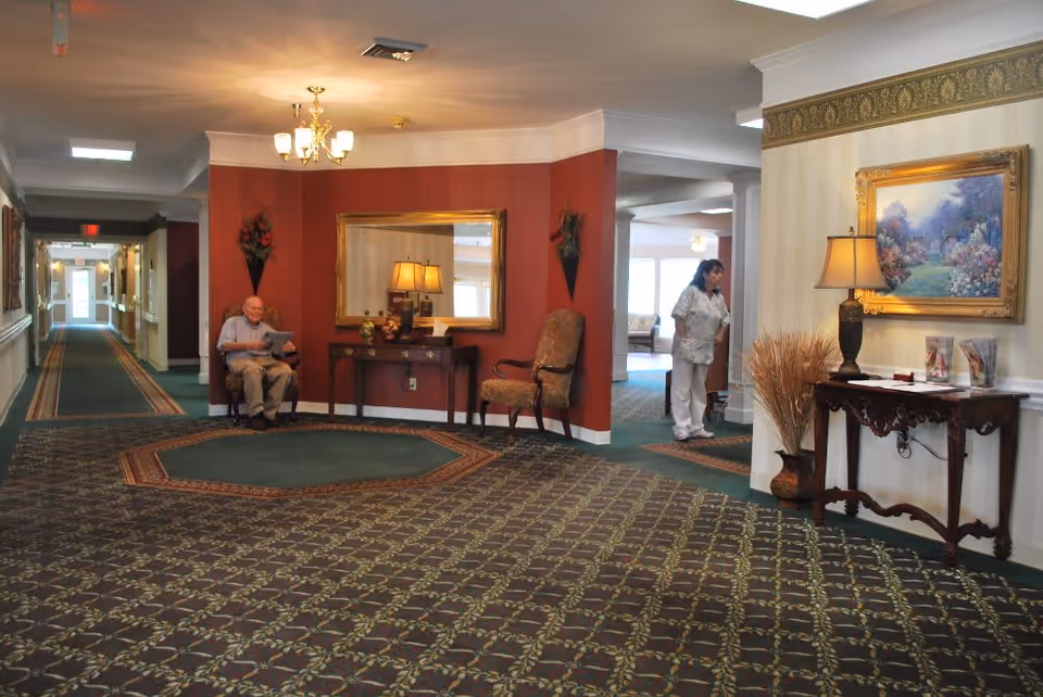 Interior view of a senior living facility hallway with patterned carpet and walls painted in red and cream. There is a man sitting on a chair reading a newspaper near a console table with a large mirror above it. A woman in a white uniform is standing in the background near another room. The hallway is decorated with framed paintings, lamps, and floral arrangements.