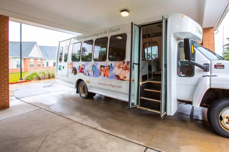 A white shuttle bus parked under a covered driveway at a senior living facility. The bus has an open door with steps leading inside and displays images of elderly people along its side. The text on the bus reads 'Retirement Living at its Finest' and includes the website www.seniorvillages.com. In the background, there is a brick building and some greenery.