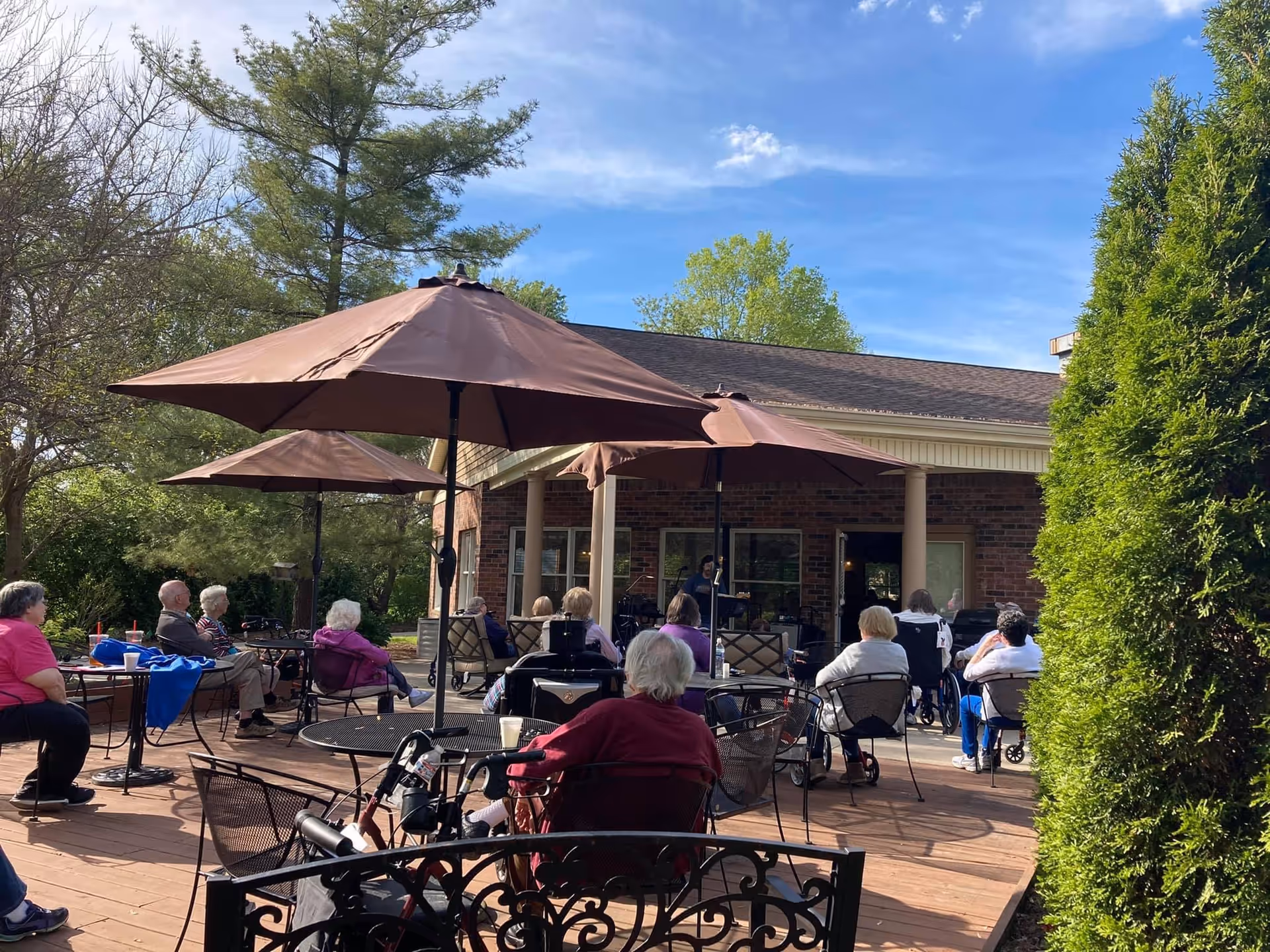 A group of elderly people sitting outdoors on a wooden deck under large brown umbrellas. They are seated in chairs and wheelchairs, facing a building with a covered porch. The scene is set on a sunny day with trees and greenery surrounding the area.