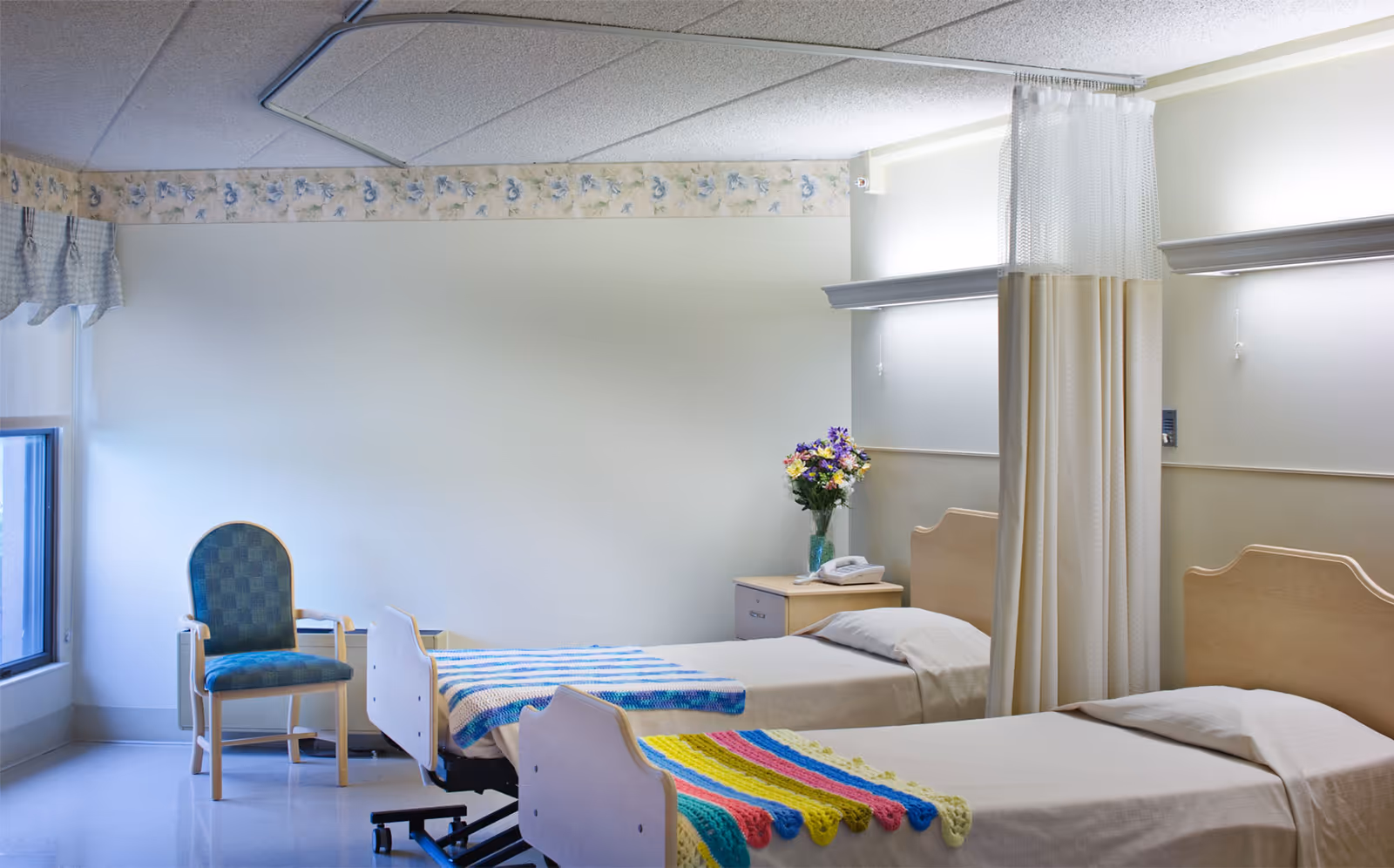 A hospital-style room with two single beds separated by a privacy curtain. Each bed has a colorful crocheted blanket at the foot. There is a nightstand with a telephone and a vase of flowers between the beds. A single upholstered chair is positioned near a window with a valance. The room has a tiled ceiling and a floral wallpaper border near the ceiling.
