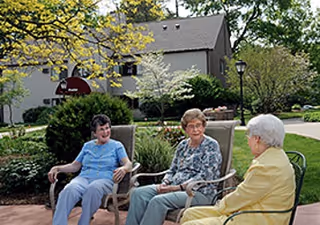 Three elderly women sitting and chatting on outdoor chairs in a garden area with trees, shrubs, and a building in the background at Day Brook Village Senior Living.