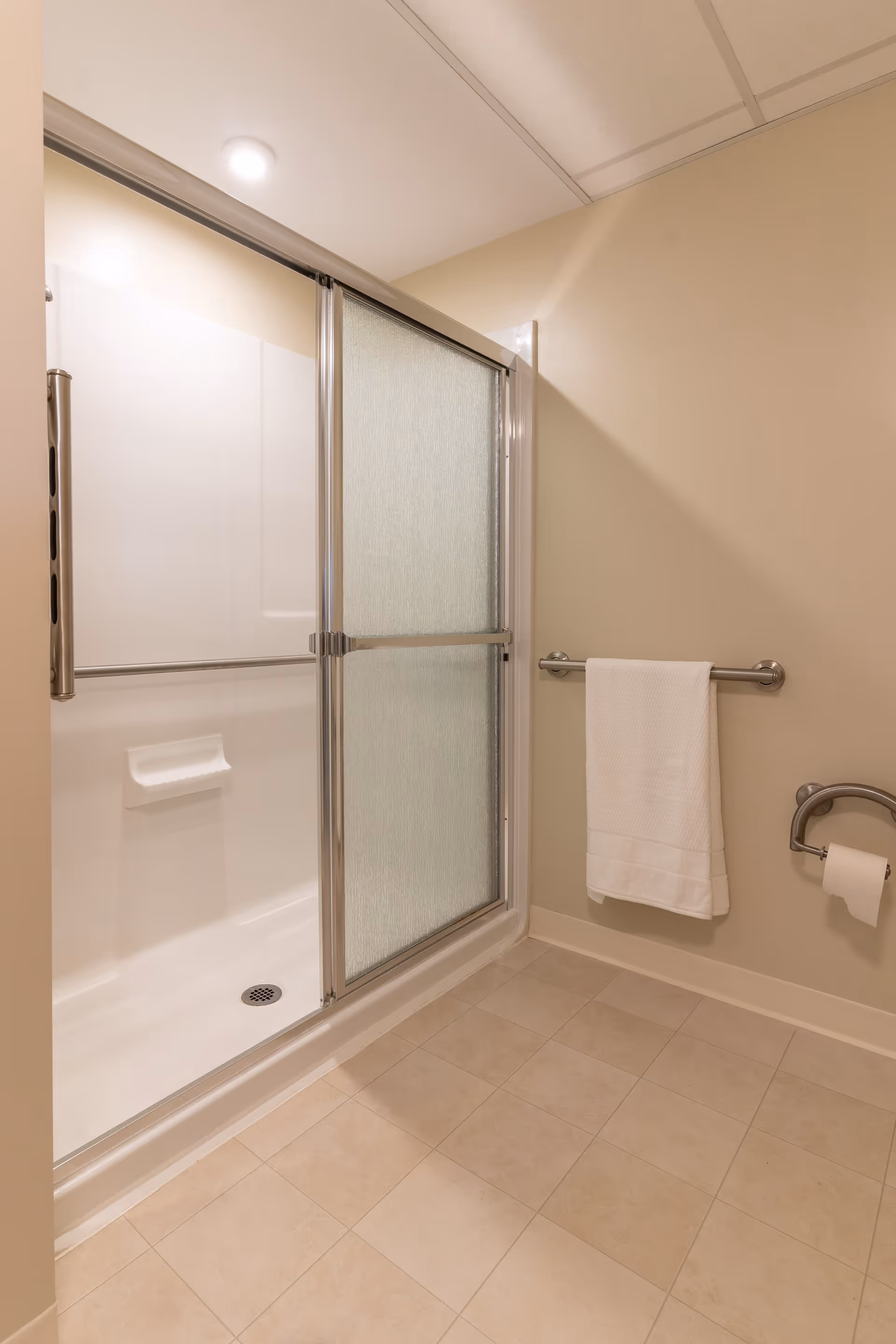 A clean bathroom with a walk-in shower featuring sliding frosted glass doors, a towel rack with a white towel, and a toilet paper holder mounted on the beige wall. The floor is tiled with light-colored tiles.