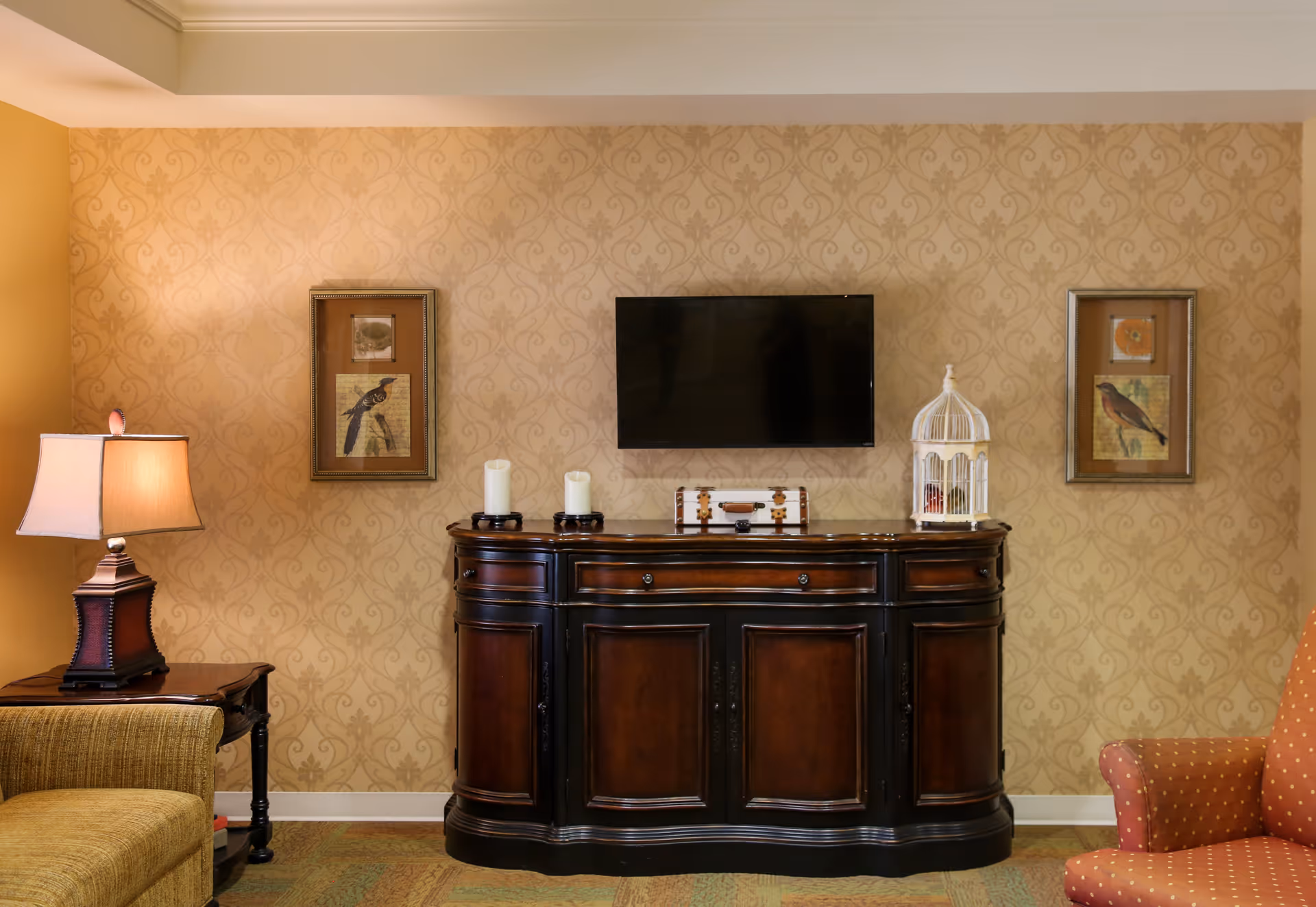 A cozy living room area with a dark wooden cabinet against a patterned beige wall. Above the cabinet is a mounted flat-screen TV. On the cabinet are two white candles, a decorative box, and a white birdcage. On either side of the TV are framed pictures of birds. To the left is a table lamp on a side table next to a beige couch, and to the right is a red armchair with a subtle pattern.