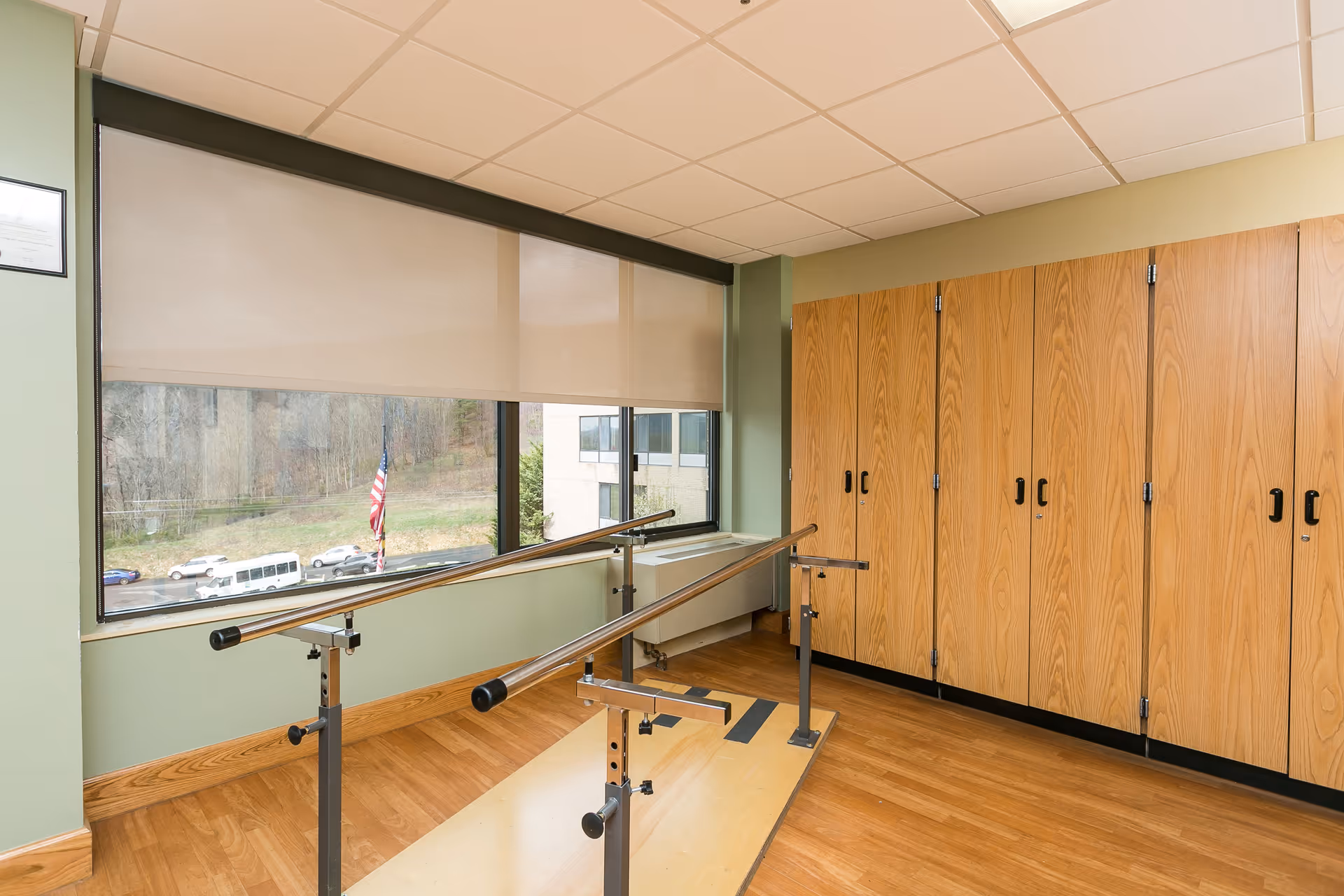 A rehabilitation room with parallel bars for physical therapy, wooden flooring, large windows with beige roller blinds, and a wall of wooden cabinets.