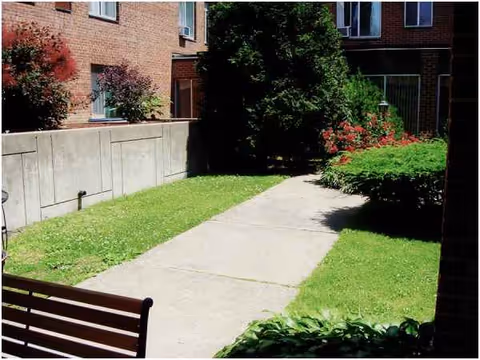A concrete walkway through a grassy outdoor courtyard beside brick buildings with benches.