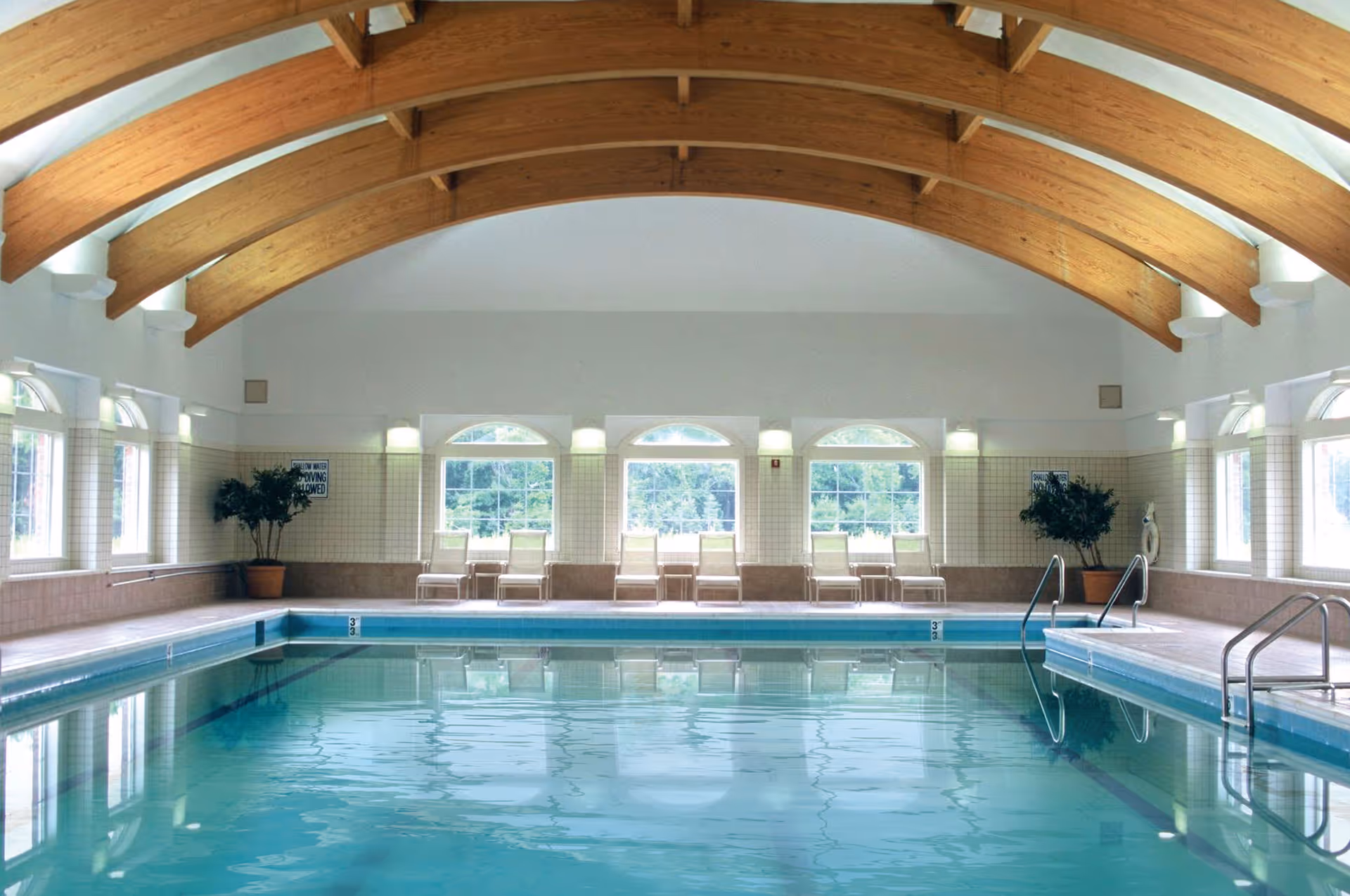 Indoor swimming pool with clear blue water, surrounded by tiled walls and large windows letting in natural light. There are several white lounge chairs lined up against the far wall, with potted plants on either side. The ceiling features wooden beams that curve gracefully across the room.