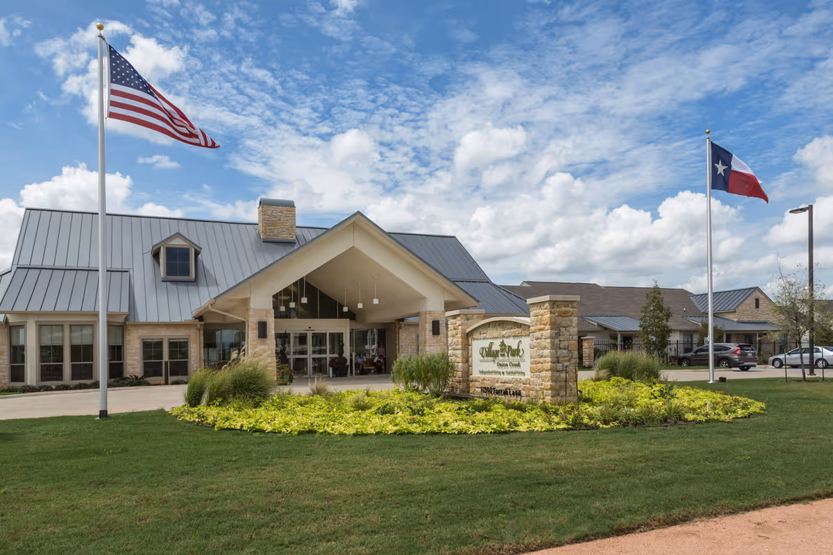 Exterior view of Village on the Park Onion Creek senior living facility with a stone entrance sign, two flagpoles displaying the American and Texas flags, a well-maintained lawn, and a partly cloudy sky.