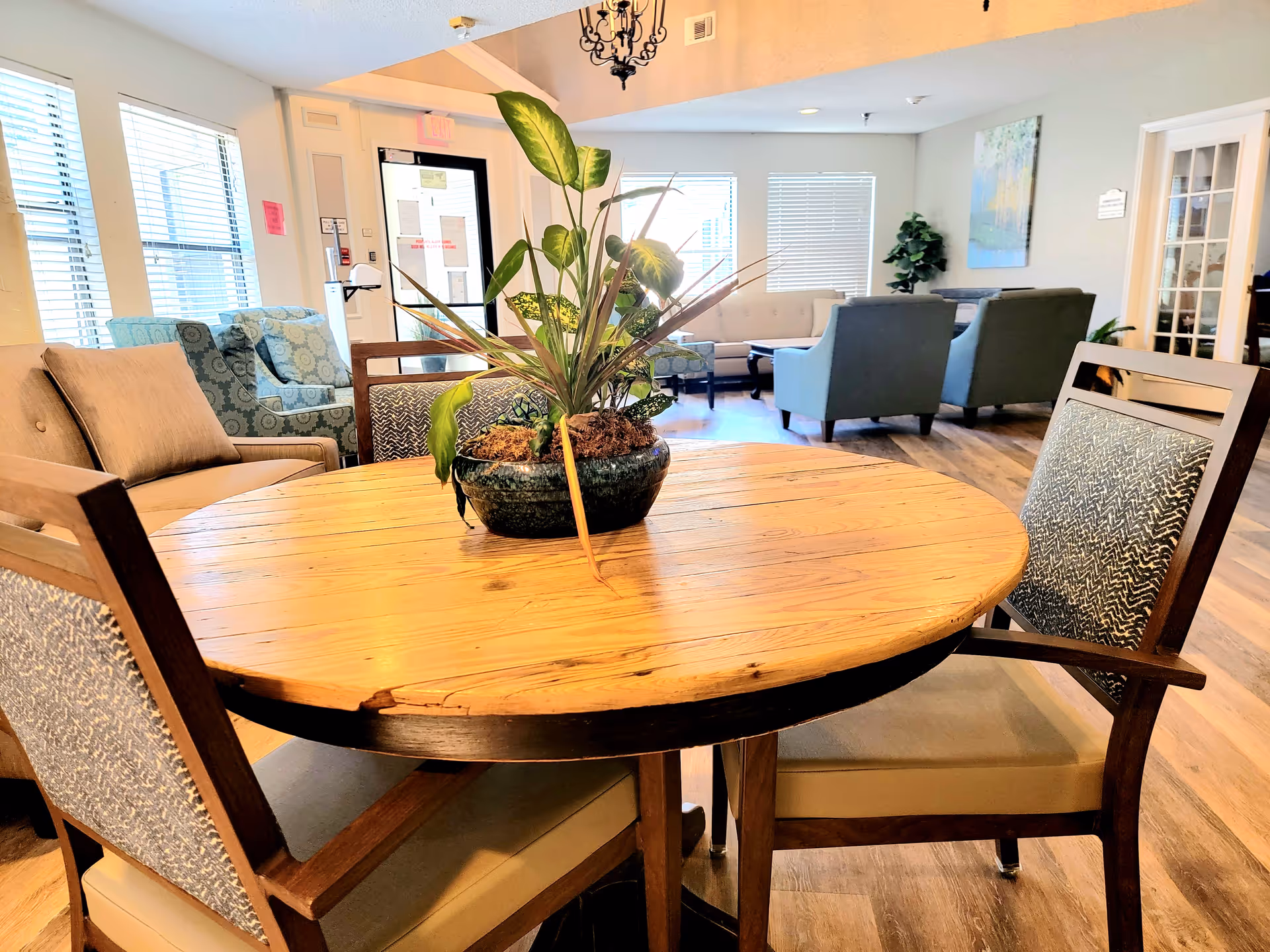 A cozy interior common area with a round wooden table in the foreground, surrounded by four cushioned chairs. A green potted plant sits in the center of the table. In the background, there are multiple upholstered chairs and sofas arranged near windows with blinds, and a door with an exit sign above it. The room has wooden flooring and soft lighting.