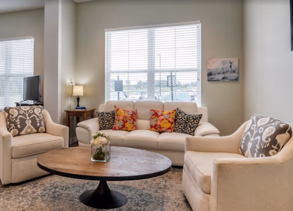 A cozy living room with a white sofa adorned with colorful floral and patterned cushions, two beige armchairs with patterned pillows, a round wooden coffee table with a small floral arrangement, a side table with a lamp, and large windows with white blinds letting in natural light.