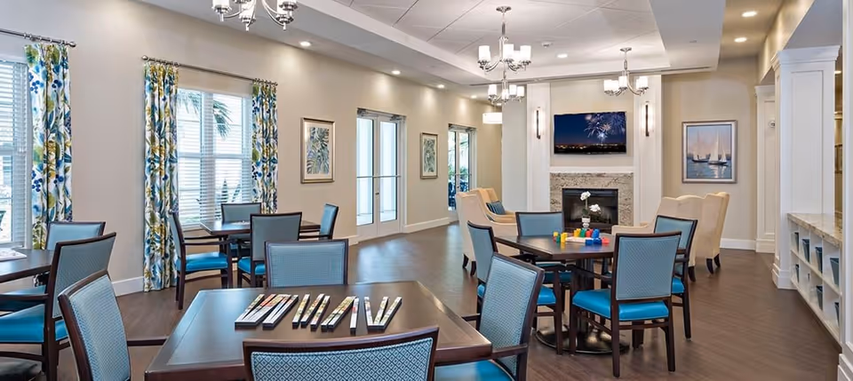 A bright and spacious common area in a senior living facility with several tables and chairs arranged for social activities. The room features large windows with floral curtains, a fireplace with a TV mounted above it, and comfortable armchairs. The walls are decorated with framed artwork, and the flooring is wood.
