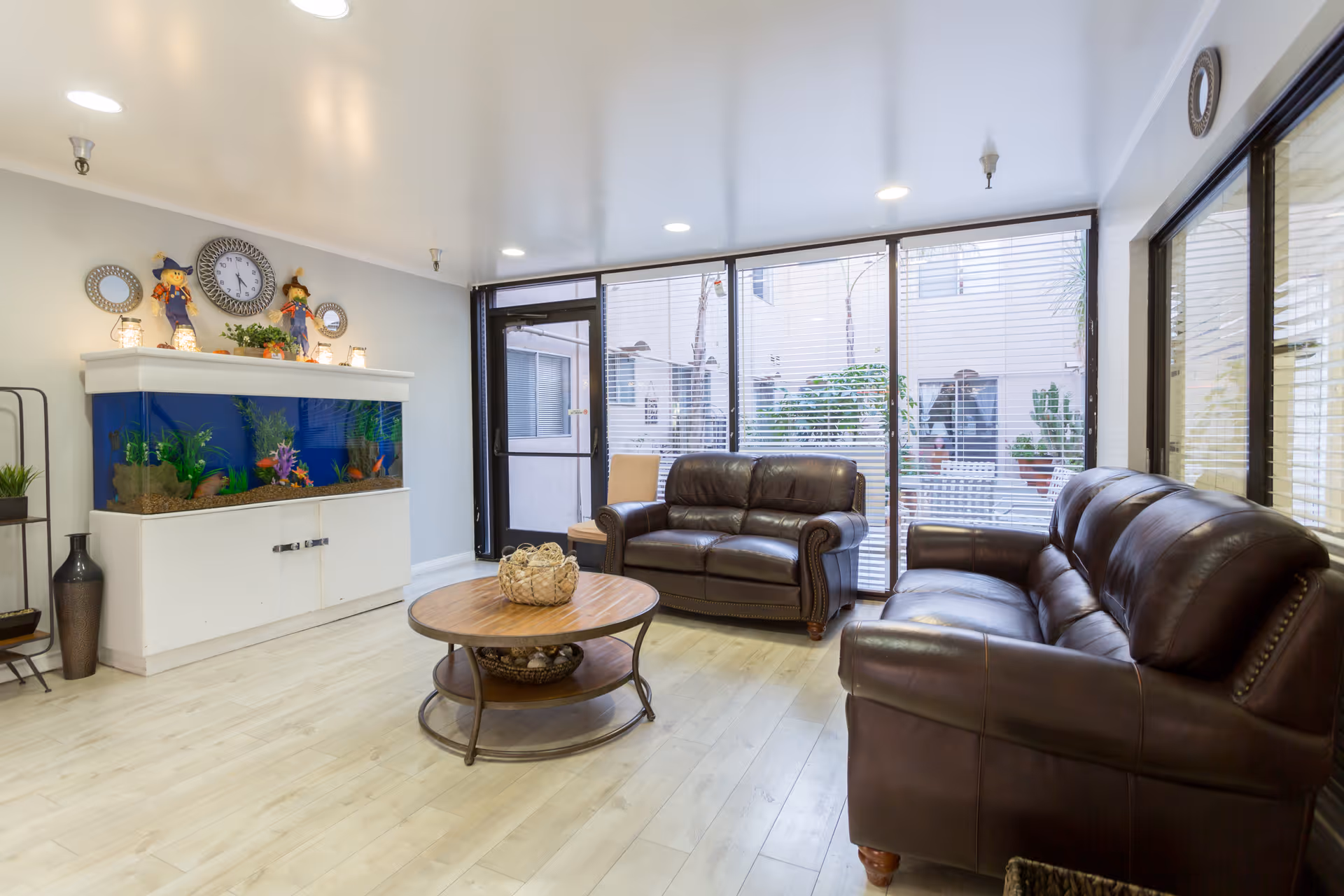 Bright common living room with brown leather sofas, a round wooden coffee table, a large aquarium, and floor-to-ceiling windows.