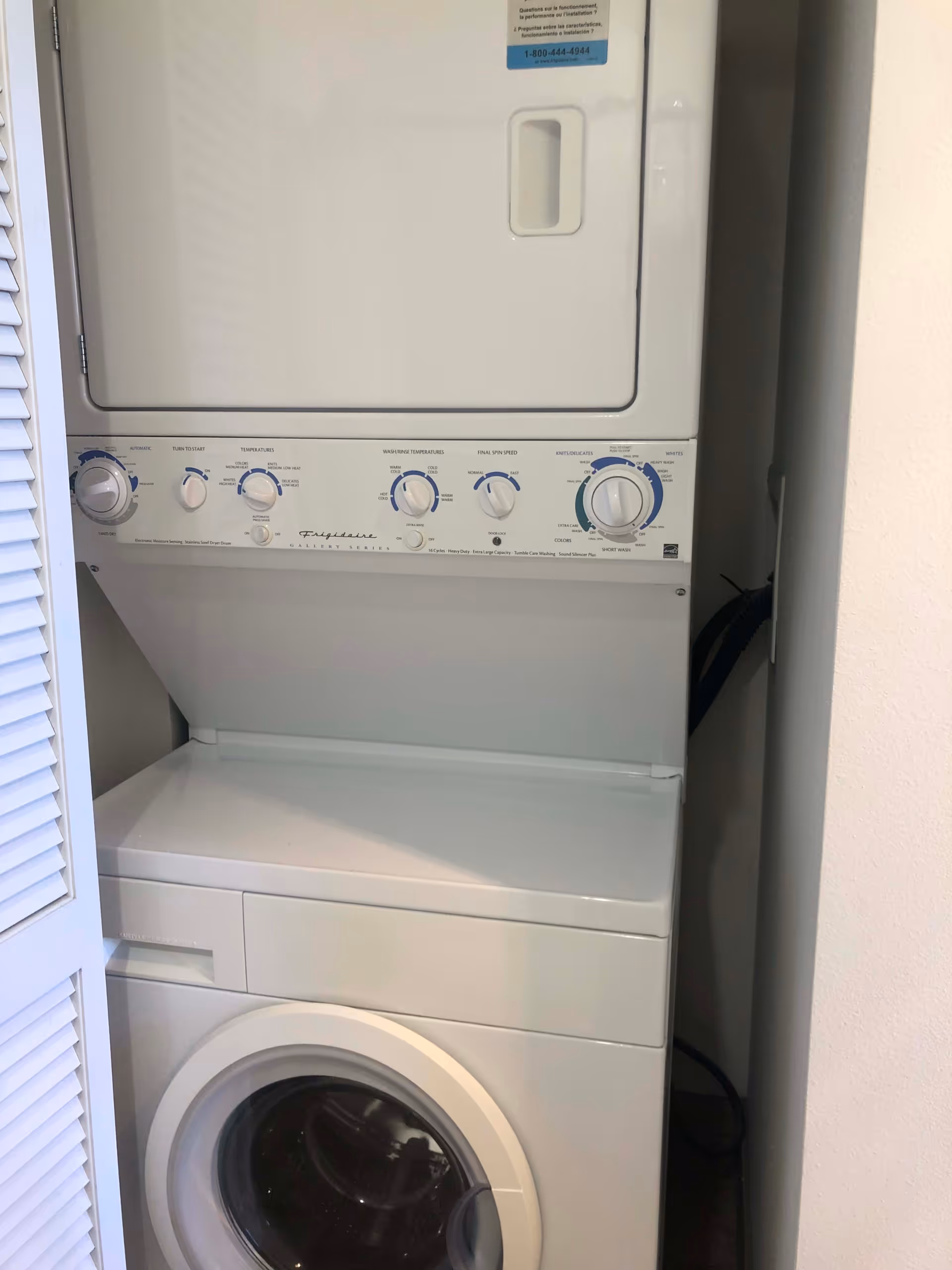 A stacked white washer and dryer unit inside a small laundry closet with a louvered door partially open.