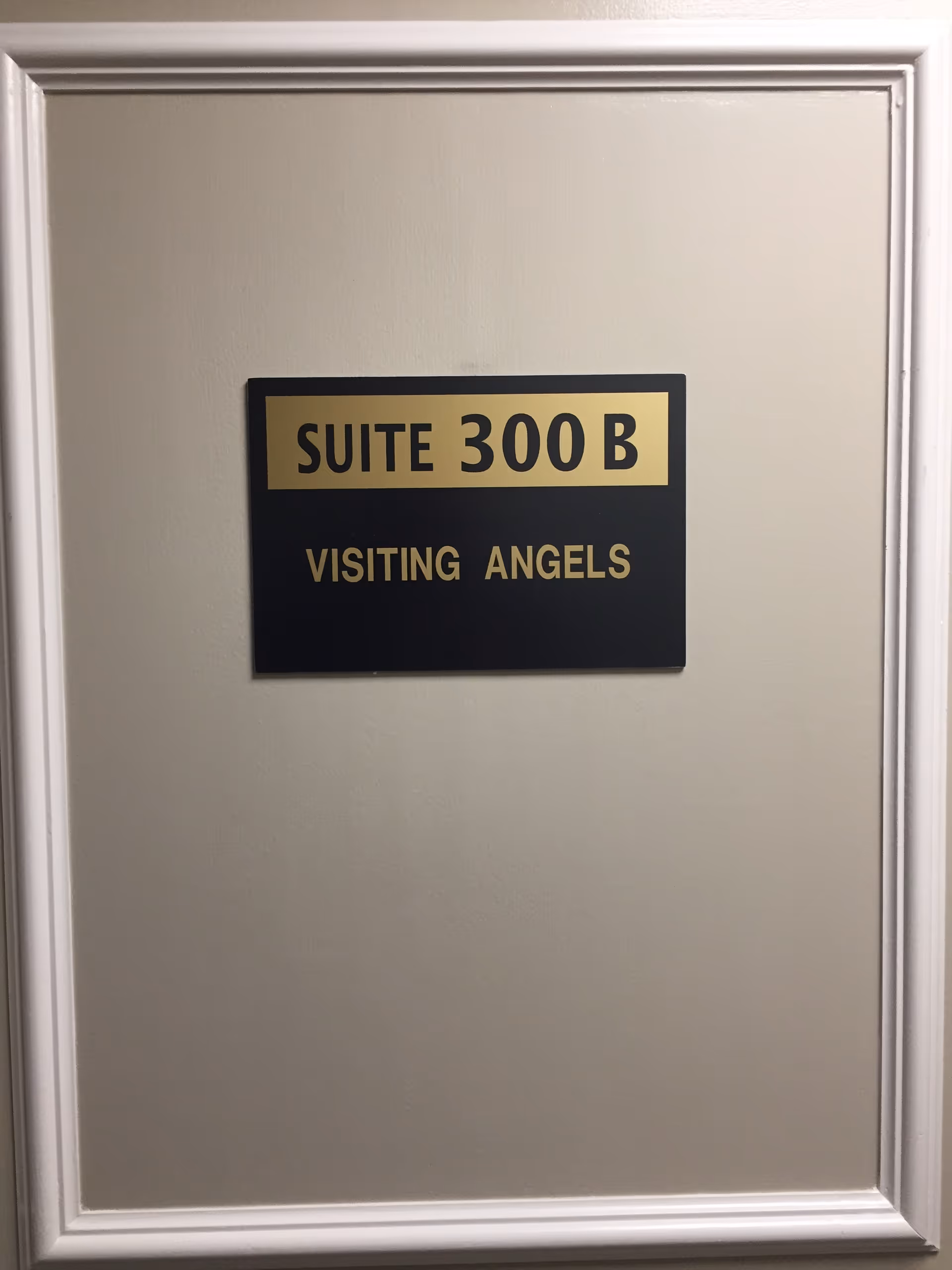 A black and gold sign mounted on a beige wall with white trim. The sign reads 'SUITE 300 B' on the top gold section and 'VISITING ANGELS' on the black section below.