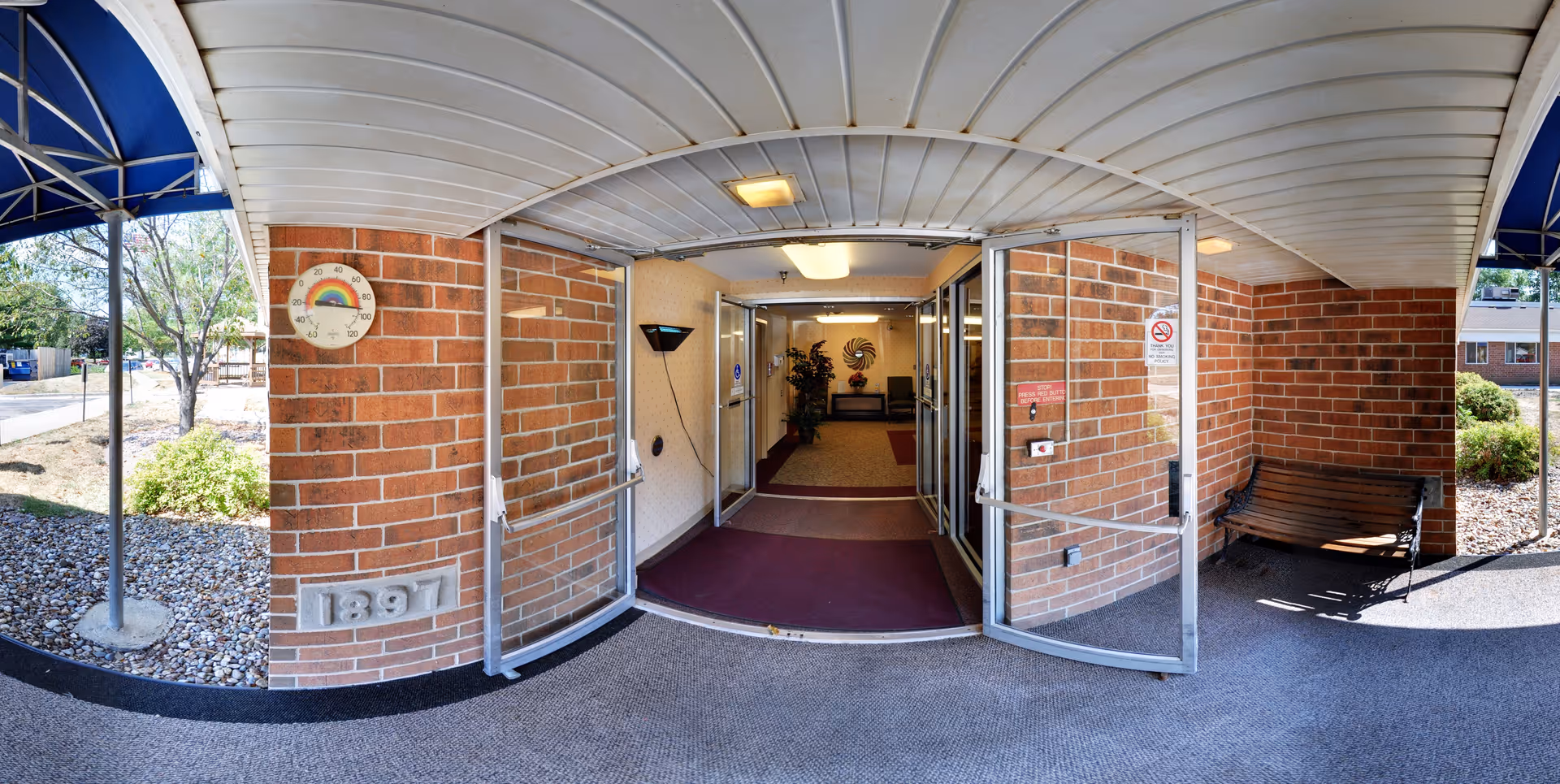 Entrance to a building with double glass doors open, showing a hallway with carpet and seating area inside. The exterior walls are made of red brick, and there is a bench on the right side outside. A thermometer with a rainbow design is mounted on the left brick wall. The area outside has a covered walkway with a blue awning and landscaping with rocks and bushes.