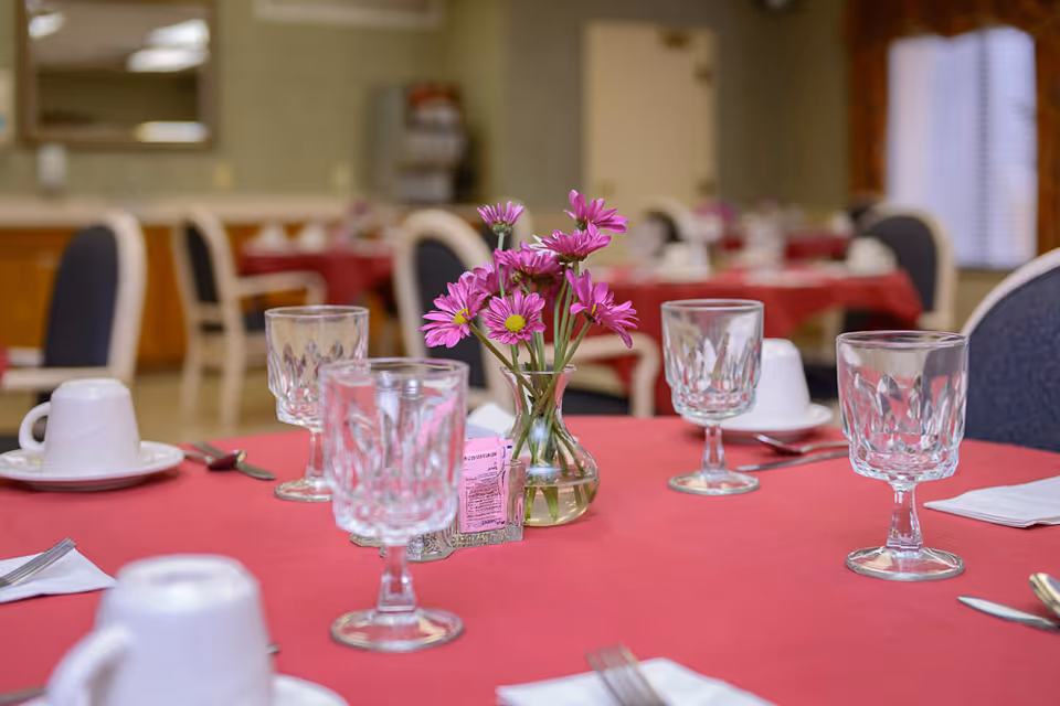 A dining table set with clear glass goblets, white cups and saucers, silverware, and a small vase with purple flowers in the center. The table is covered with a red tablecloth and other similarly set tables and chairs are visible in the softly focused background.