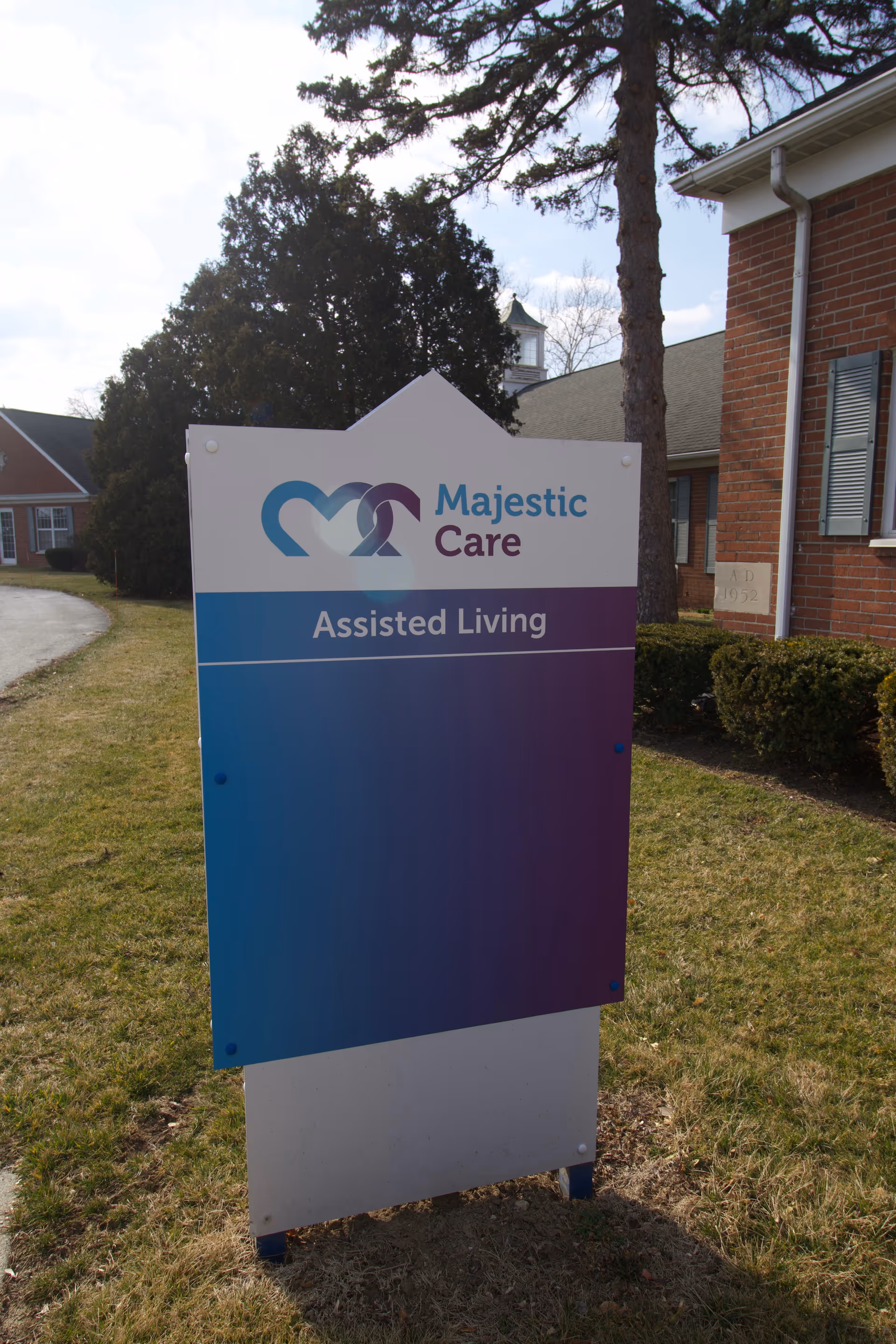 Outdoor sign for Majestic Care Assisted Living facility standing on grass near a brick building with trees and a driveway in the background.